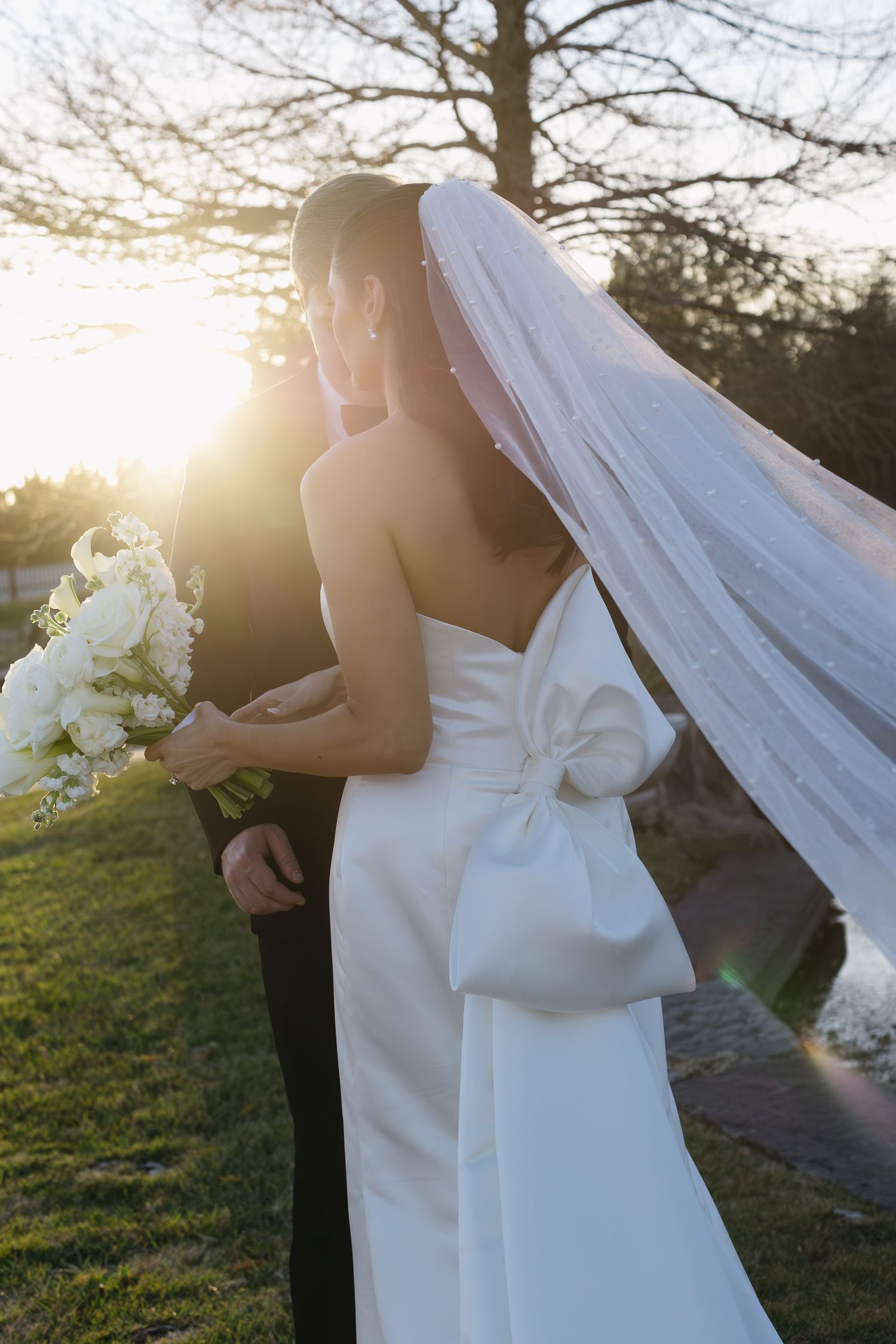 Bride in elegant white gown with bow detail holds bouquet during sunset wedding portrait.