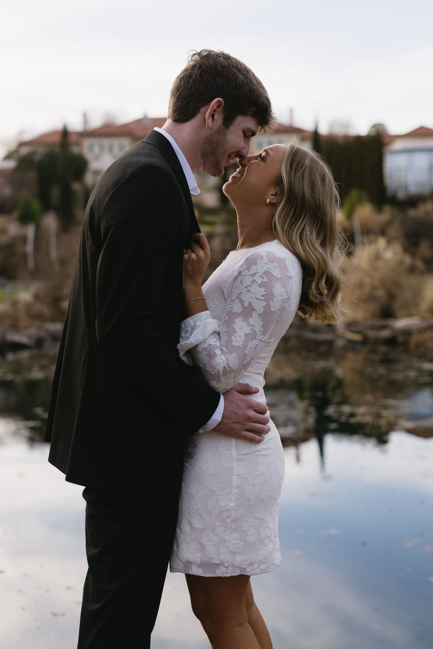 A couple embraces by a reflective lake with Mediterranean buildings in the background.