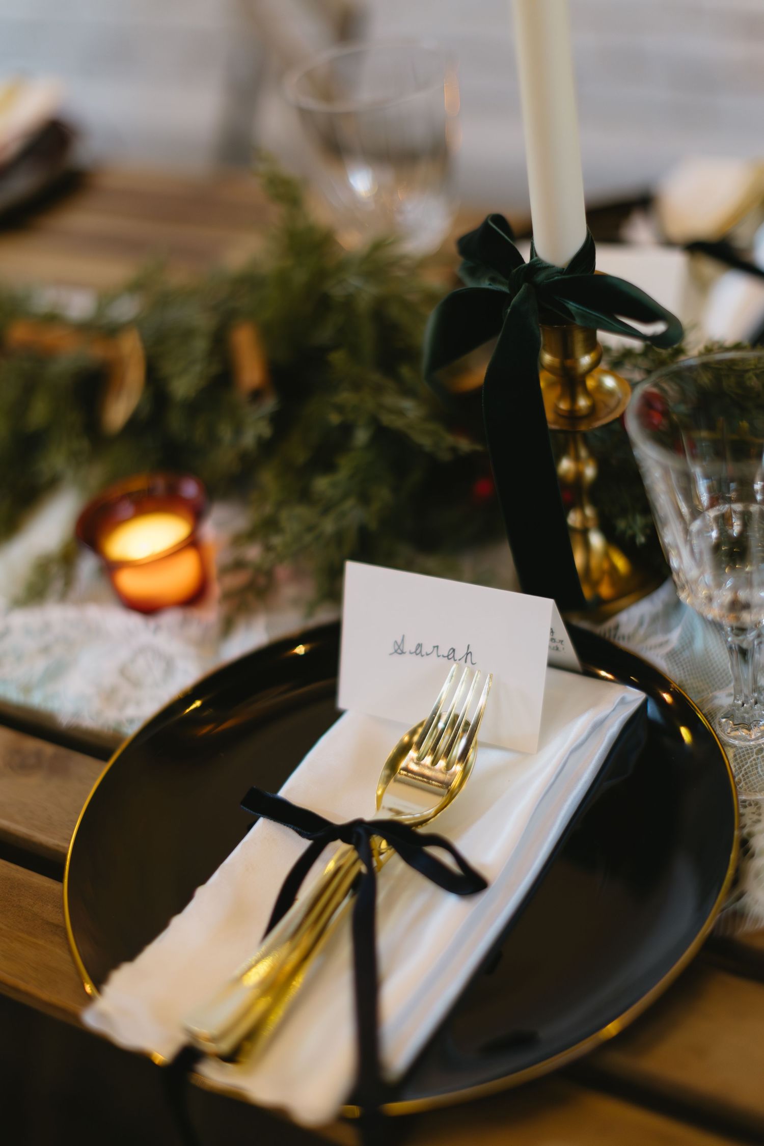 Elegant table setting with black plate, gold cutlery, white napkin and candle with evergreen decor on wooden surface.
