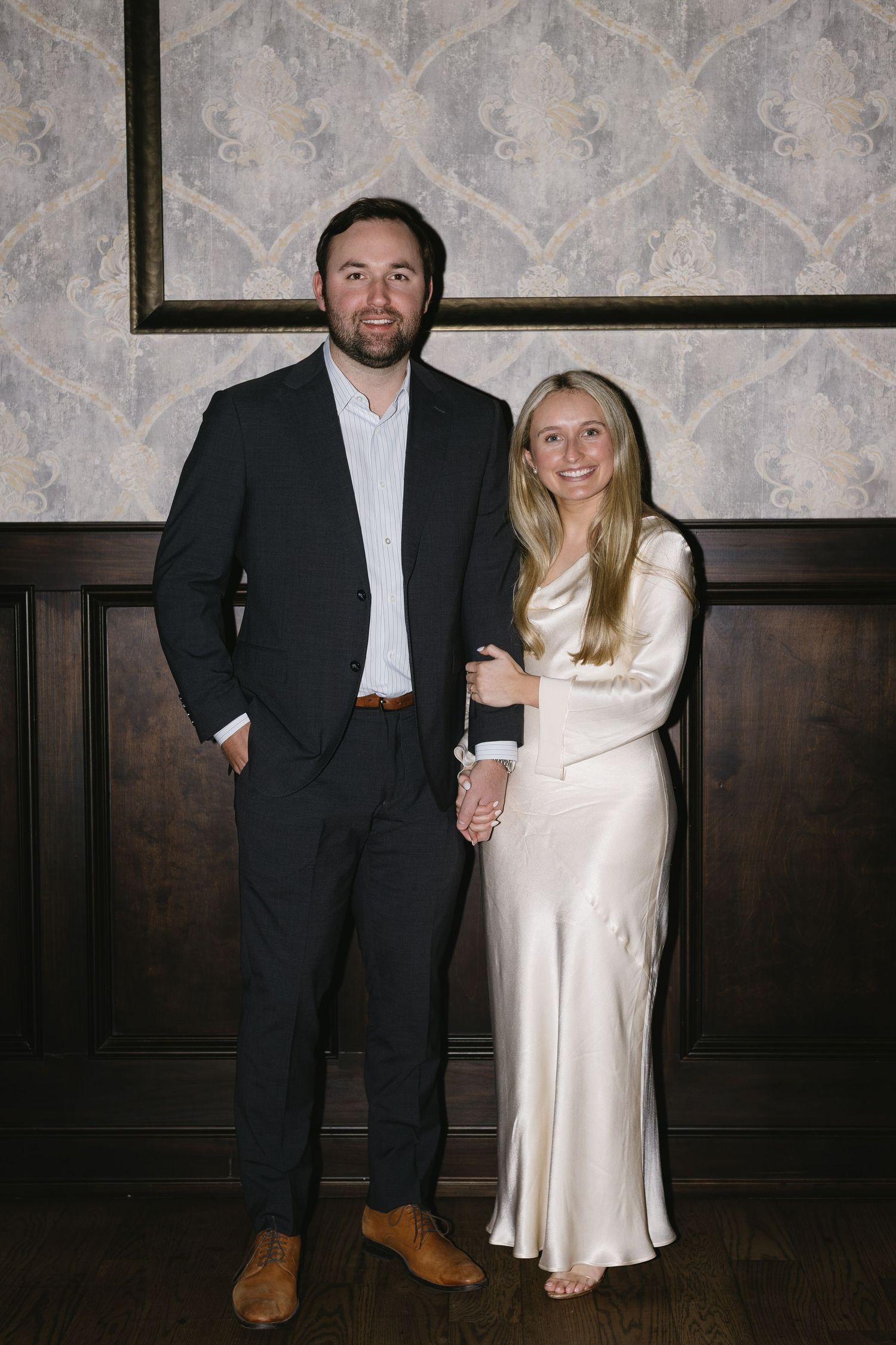 Couple in formal attire posing together against dark wood paneled wall at an event.