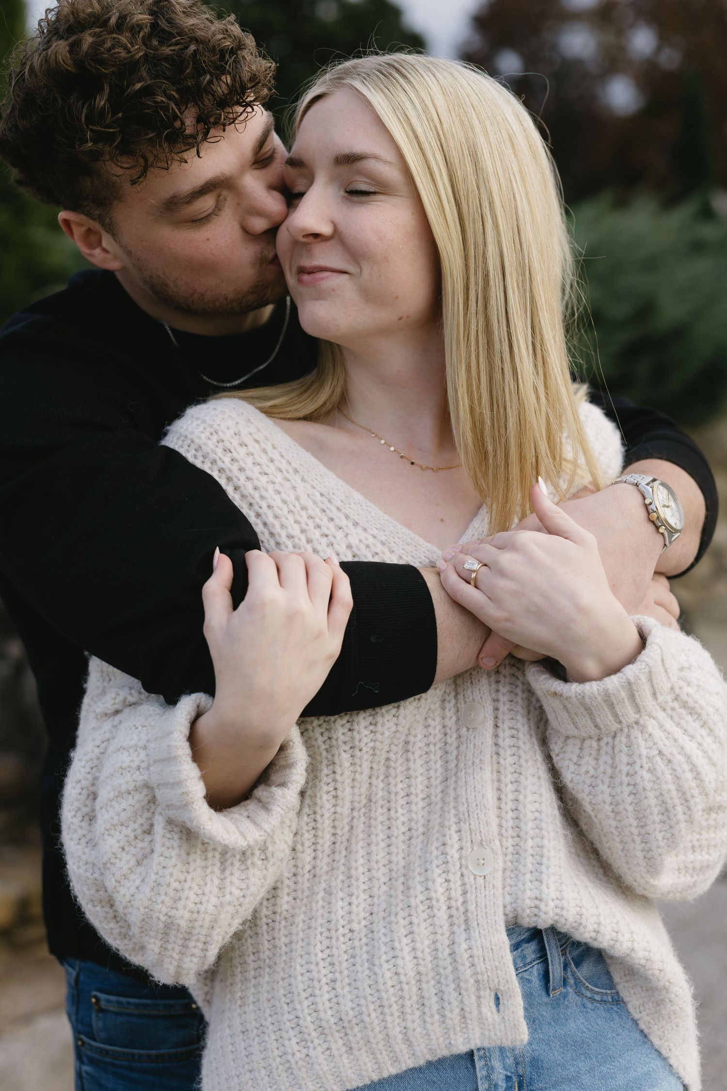 A couple shares an intimate moment while wearing a white knit sweater and black top against a blurred outdoor background.