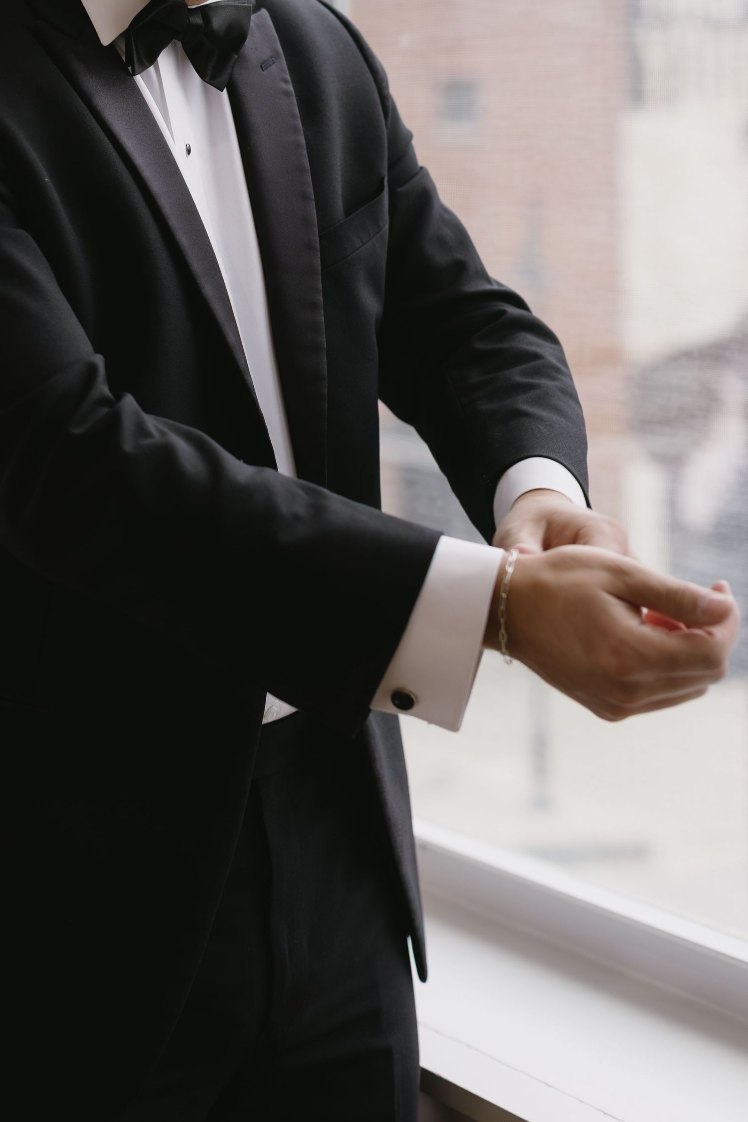 Close-up view of hands adjusting cufflinks on a formal black tuxedo sleeve near a window.