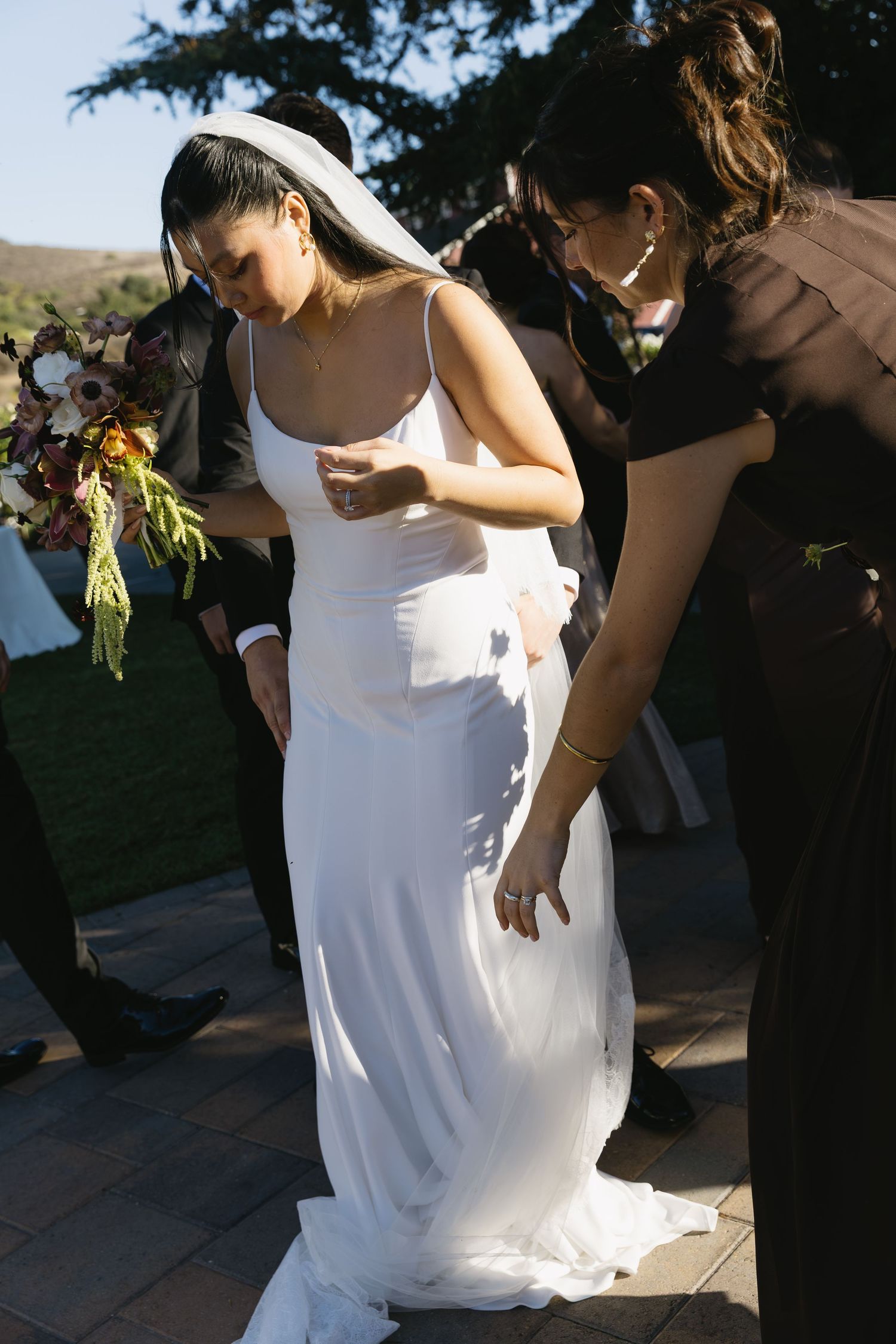 Wedding party members assist with the bride's dress during outdoor photos.