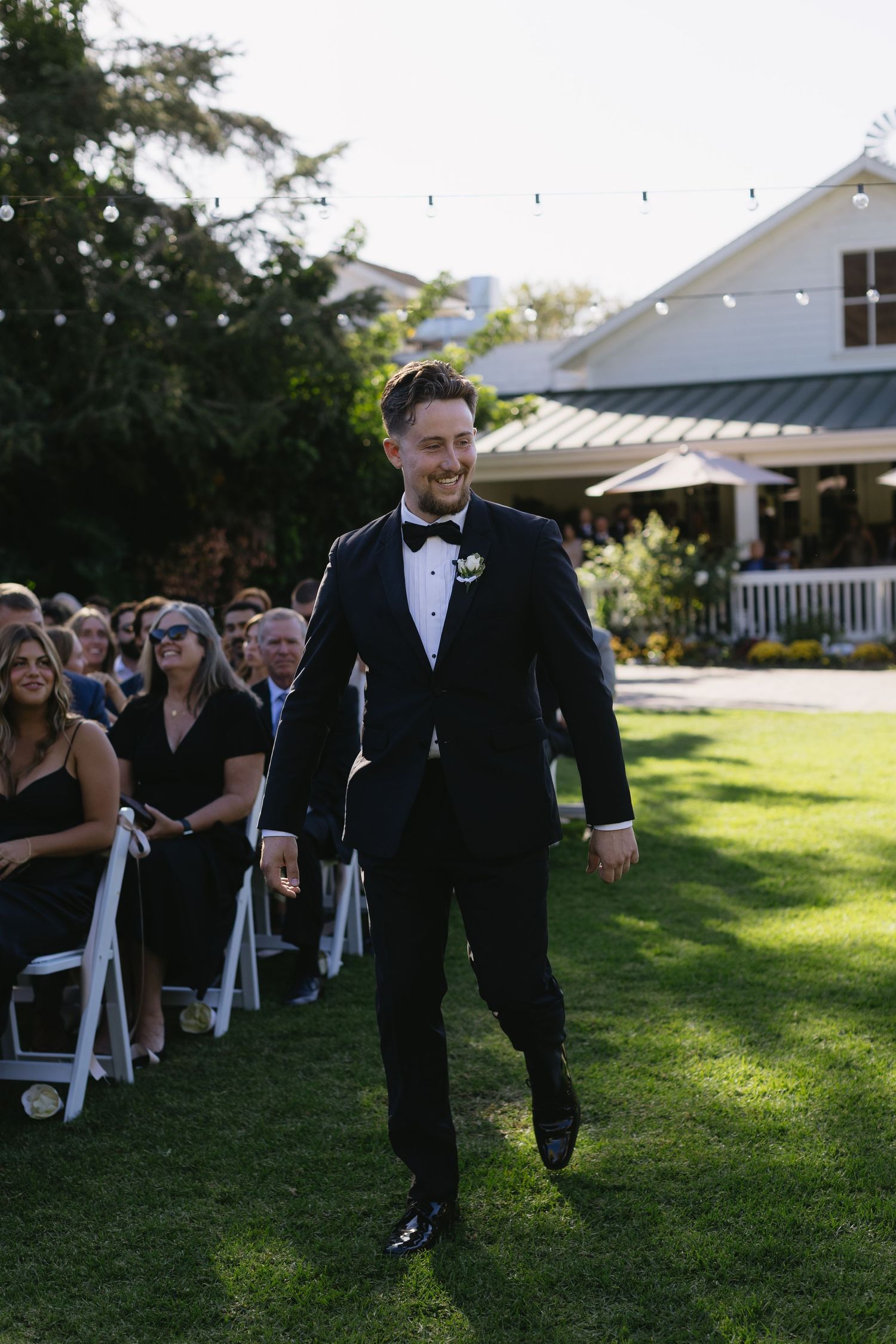A groomsman in a black tuxedo walks down an outdoor aisle with white chairs and a house in the background.