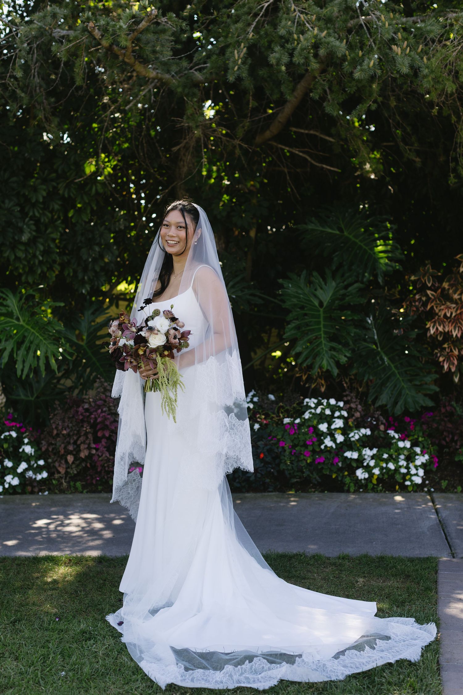 A bride in an elegant white wedding dress poses in a lush garden setting with trailing veil and floral bouquet.