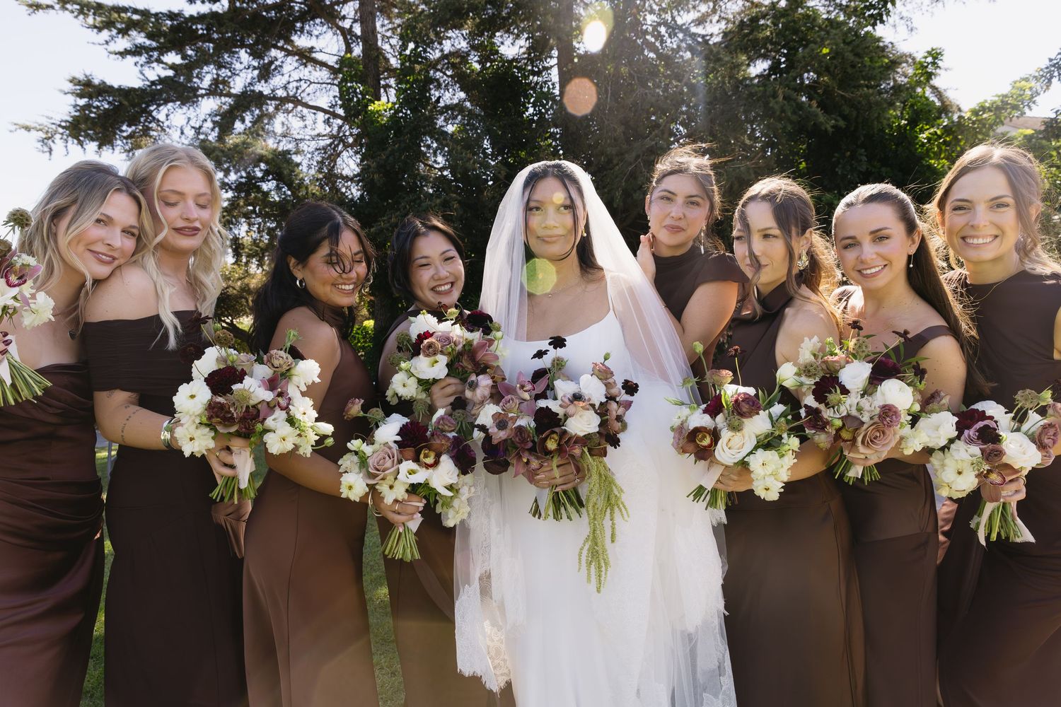 Wedding party in brown dresses posing with floral bouquets outdoors.