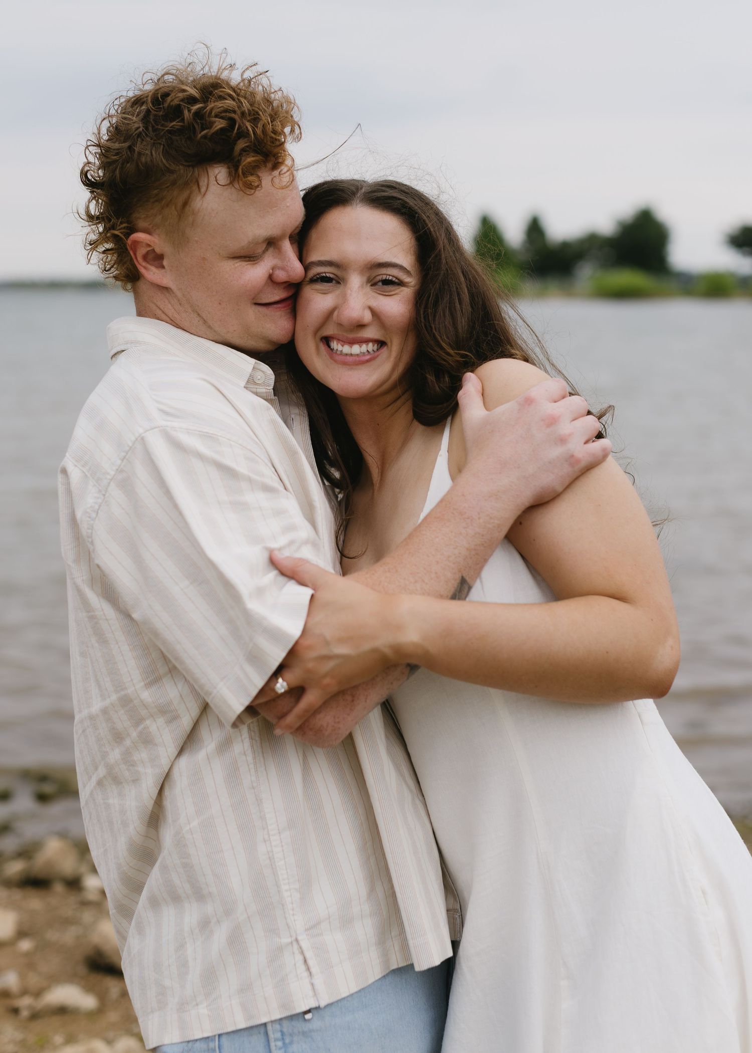 A couple embraces lovingly by the water on an overcast day.