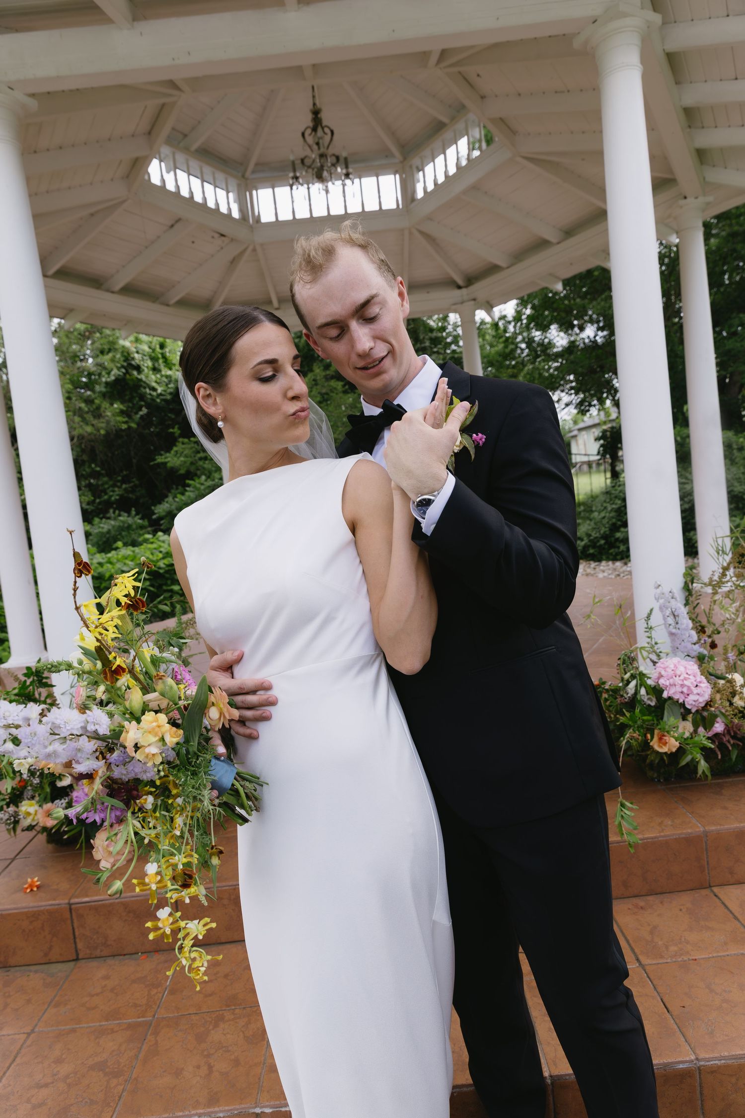 A couple shares a romantic dance moment under an elegant white gazebo decorated with colorful floral arrangements.