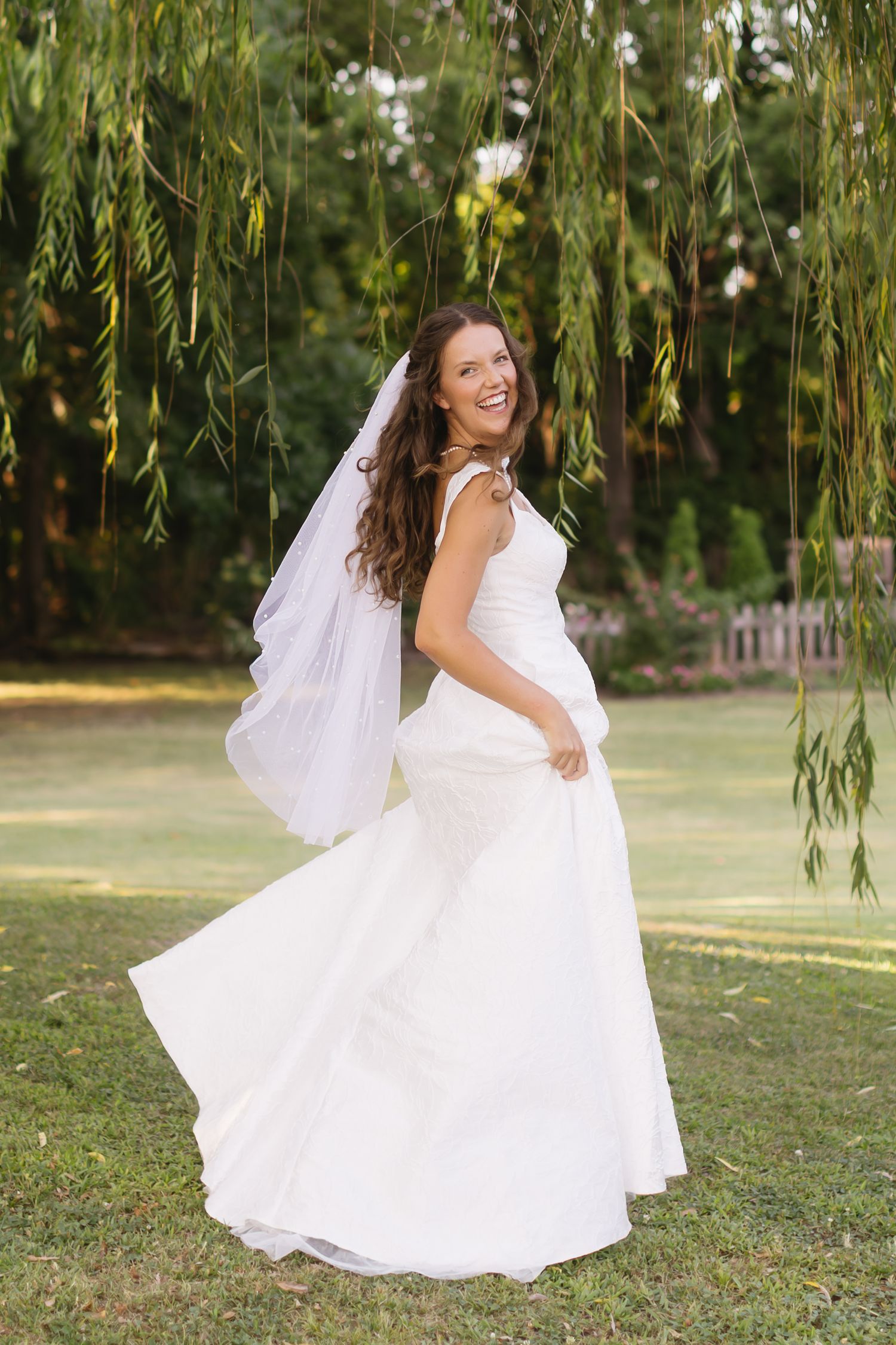 A bride spins gracefully in her flowing white gown under weeping willow branches.