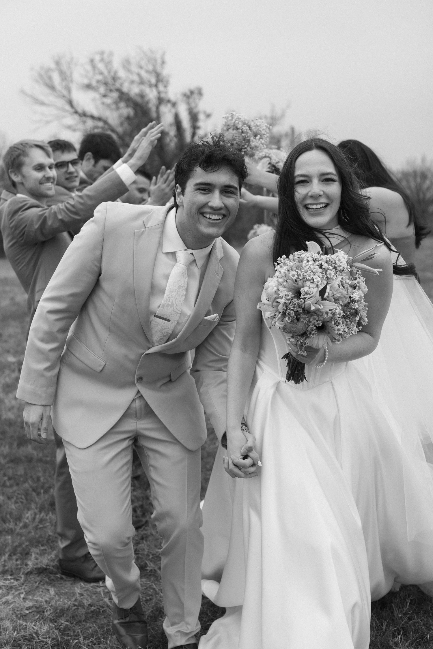 A joyful wedding couple celebrates with guests cheering behind them in a black and white photograph.