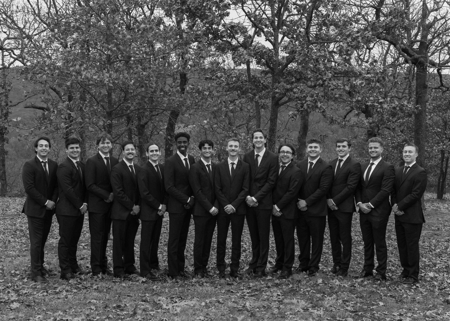 Large wedding party in formal black attire lined up for group photo in autumn setting.