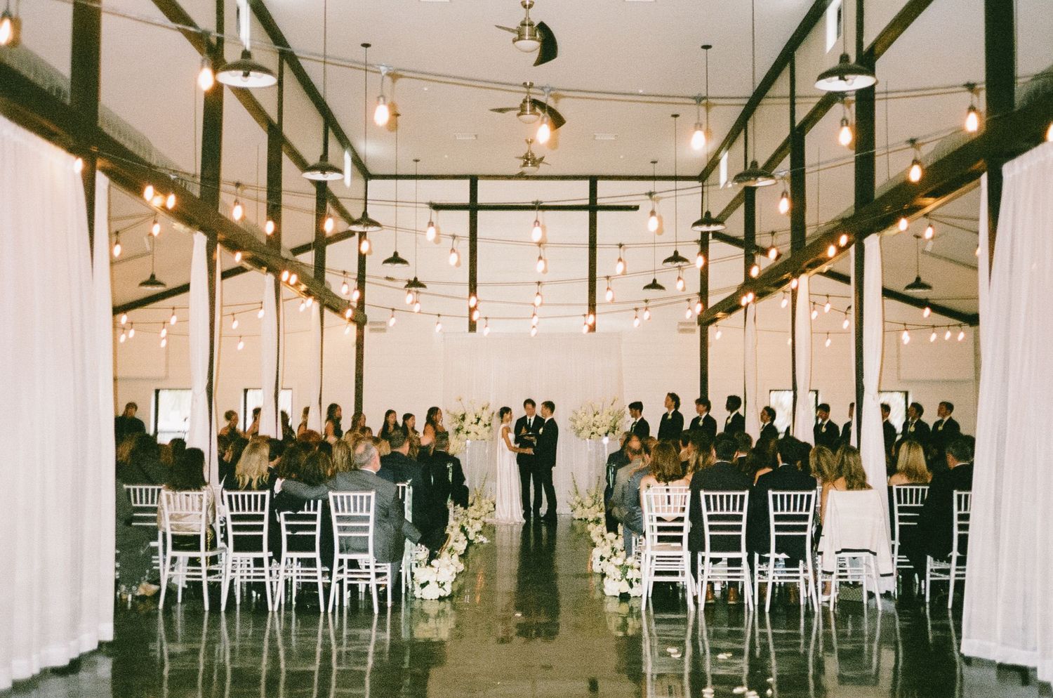 Modern industrial wedding ceremony space with white drapes and string lights overhead.