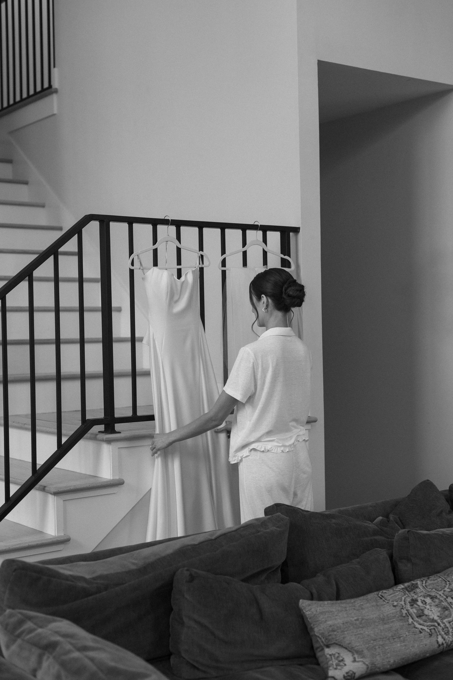 A person in white attends to a hanging dress near a staircase in a monochrome interior.
