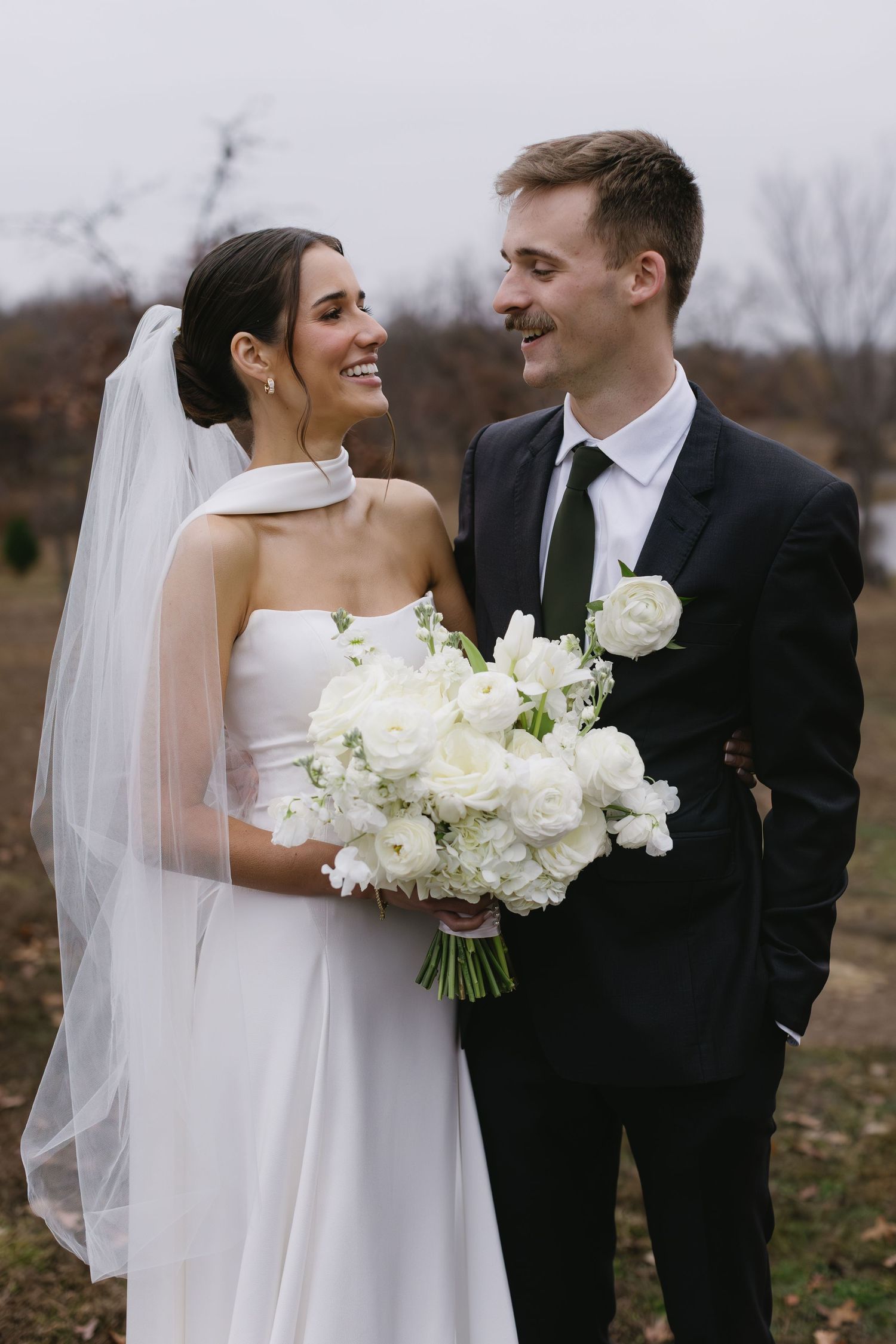 Newlyweds posing with white floral bouquet against autumn landscape.