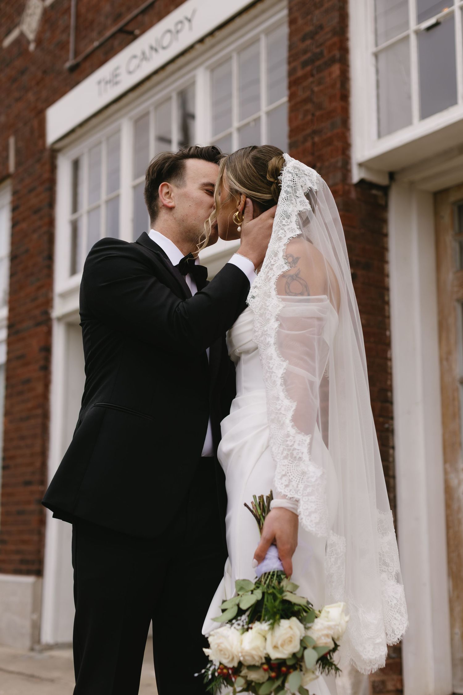 A romantic wedding kiss captured outside a classic brick building with white trim.