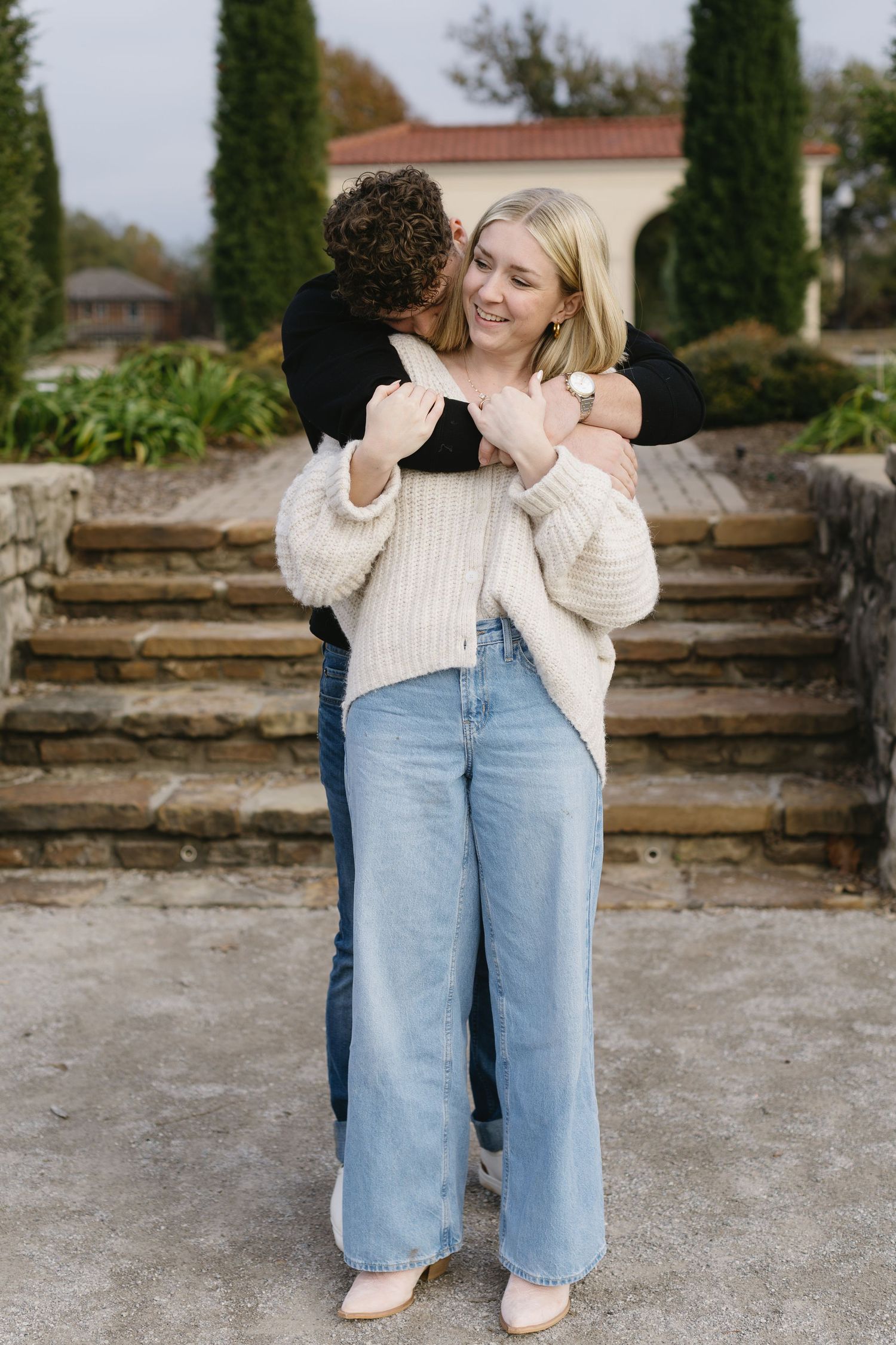 Two people in light sweaters and blue jeans stand together on a stone patio near tall cypress trees.