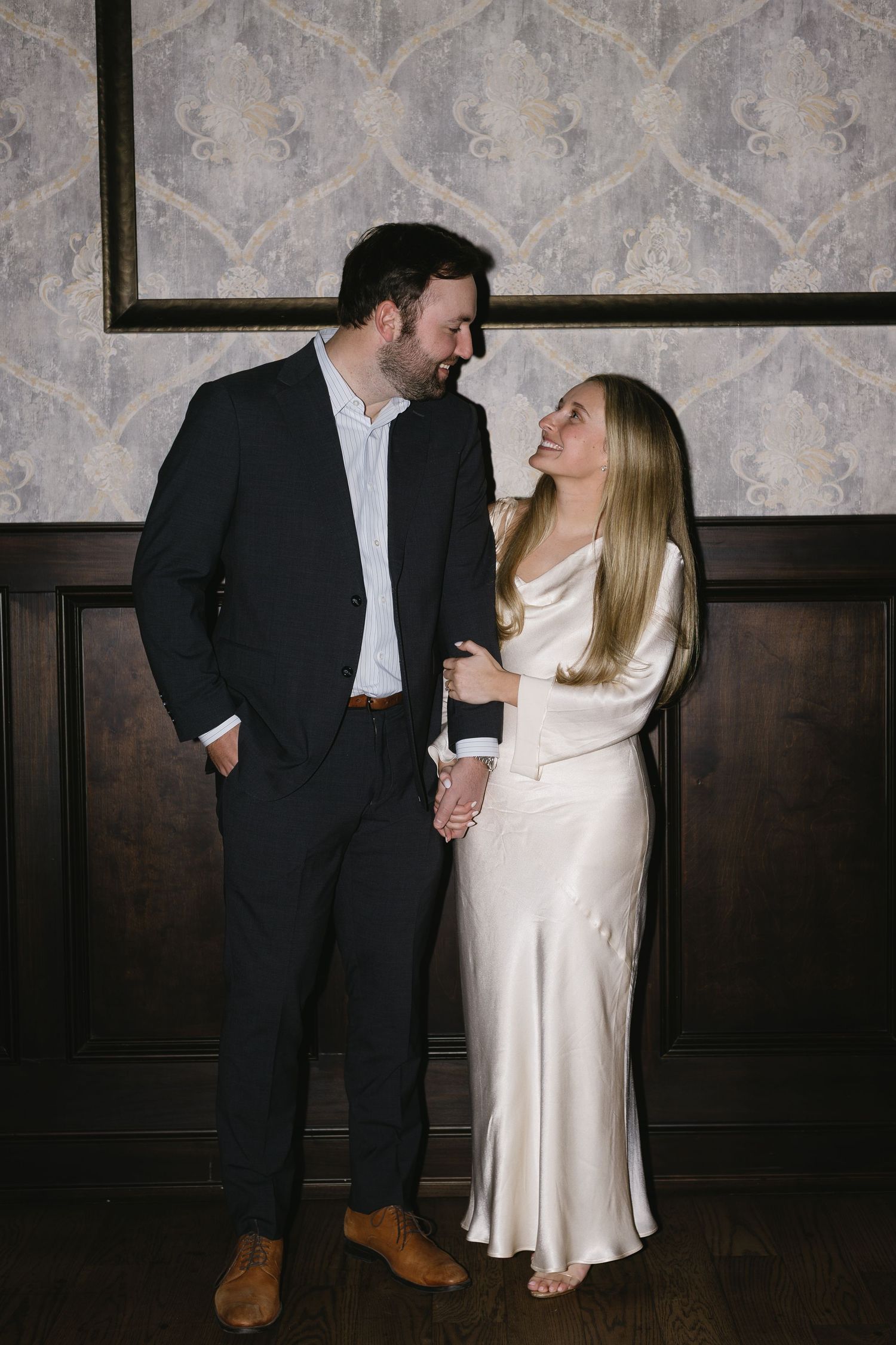 A couple shares an intimate moment in formal attire against a patterned wallpaper background.