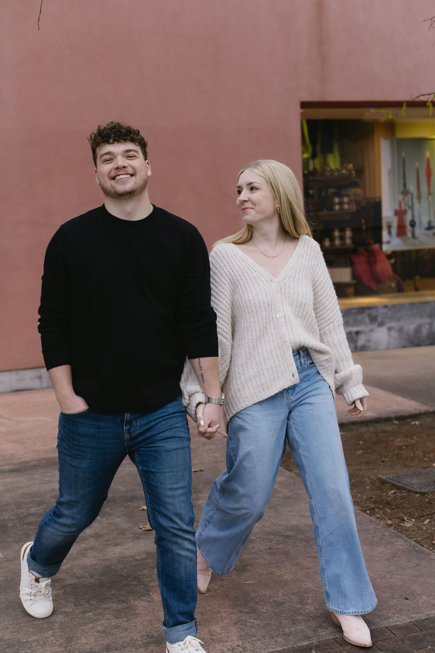 A couple walking playfully together against a pink wall wearing coordinated casual outfits.