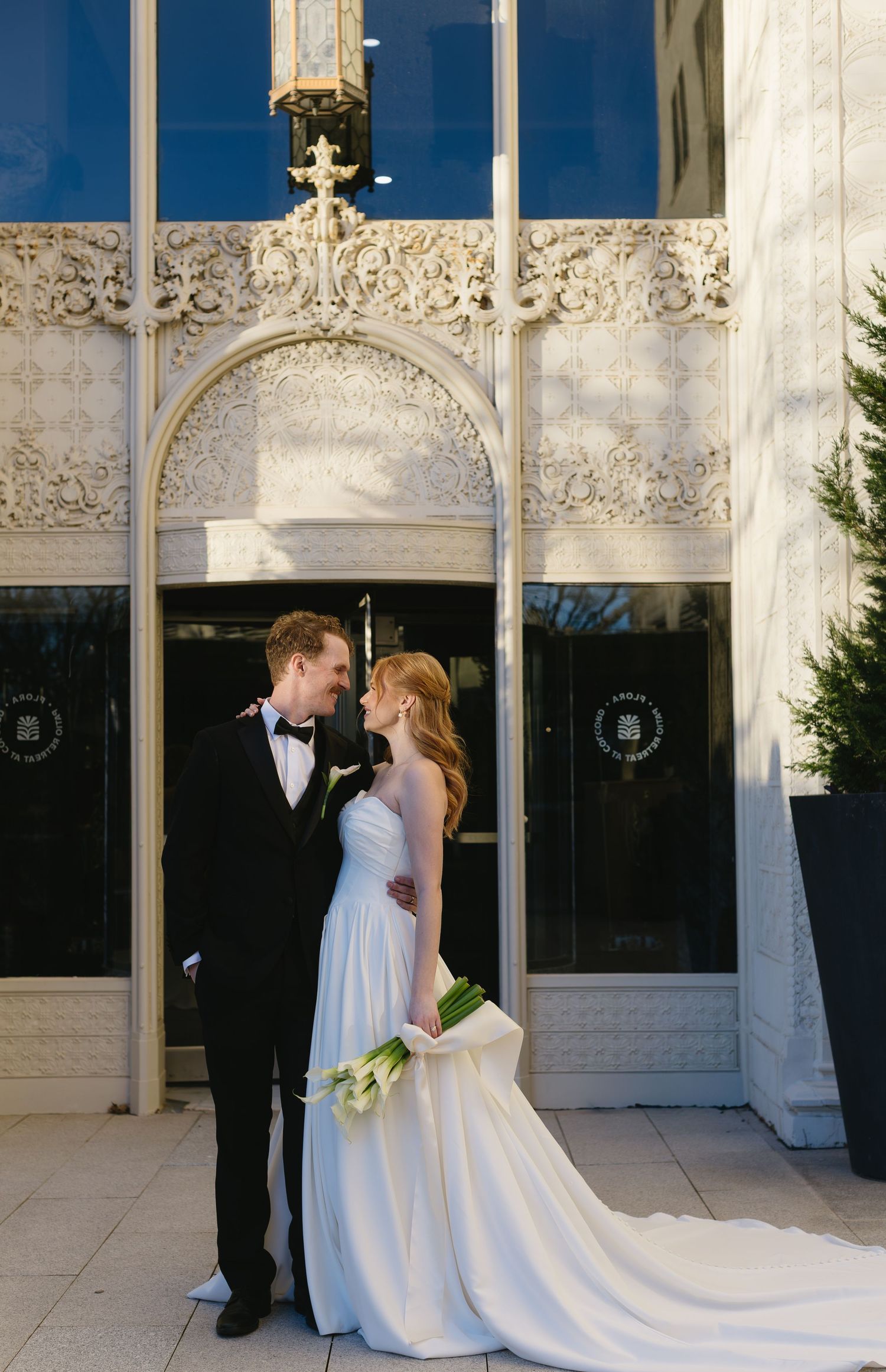 Newlyweds pose beneath an ornate white architectural arch with elegant carvings.
