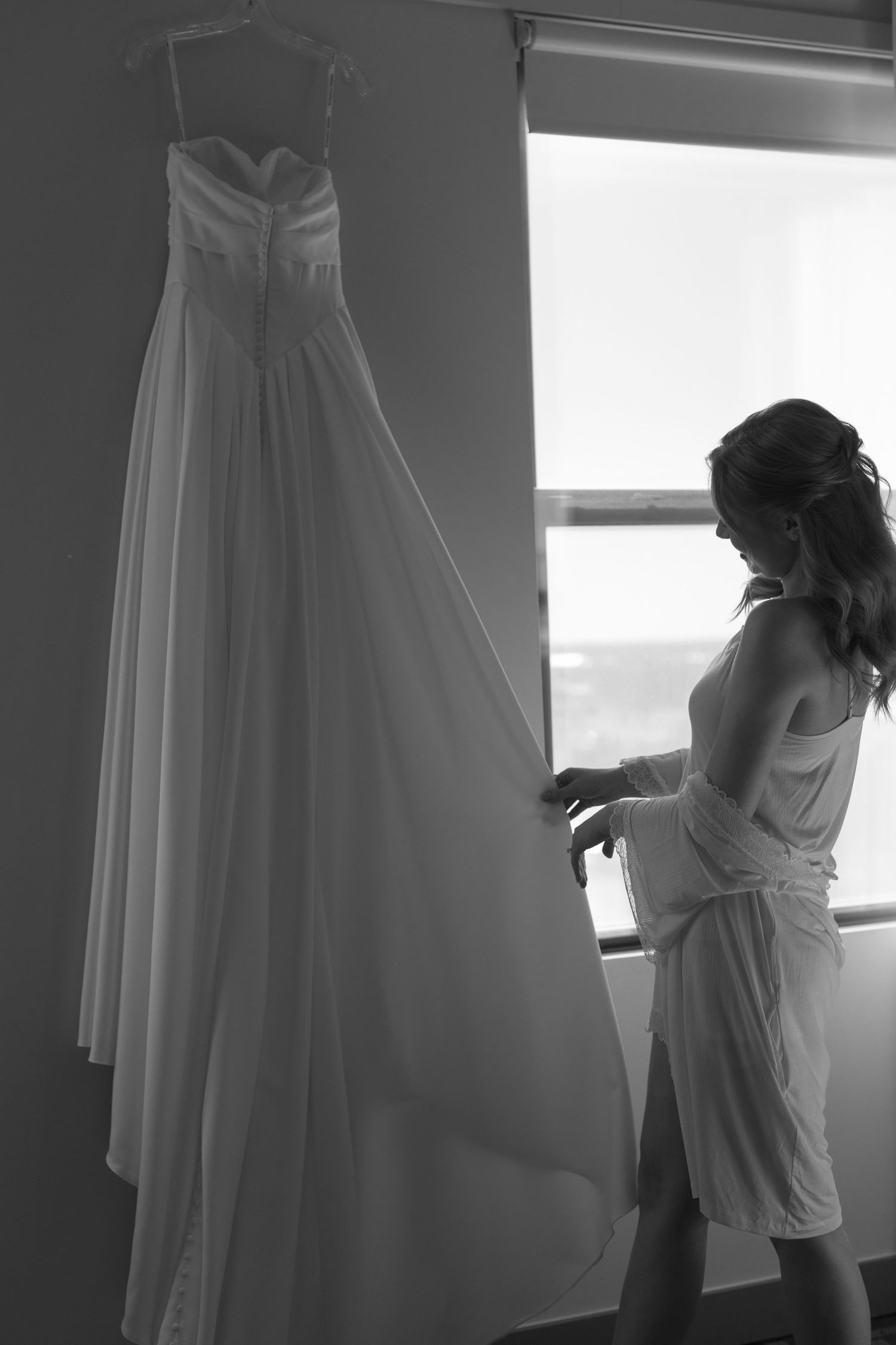 Silhouettes preparing wedding dress by window in black and white photography.