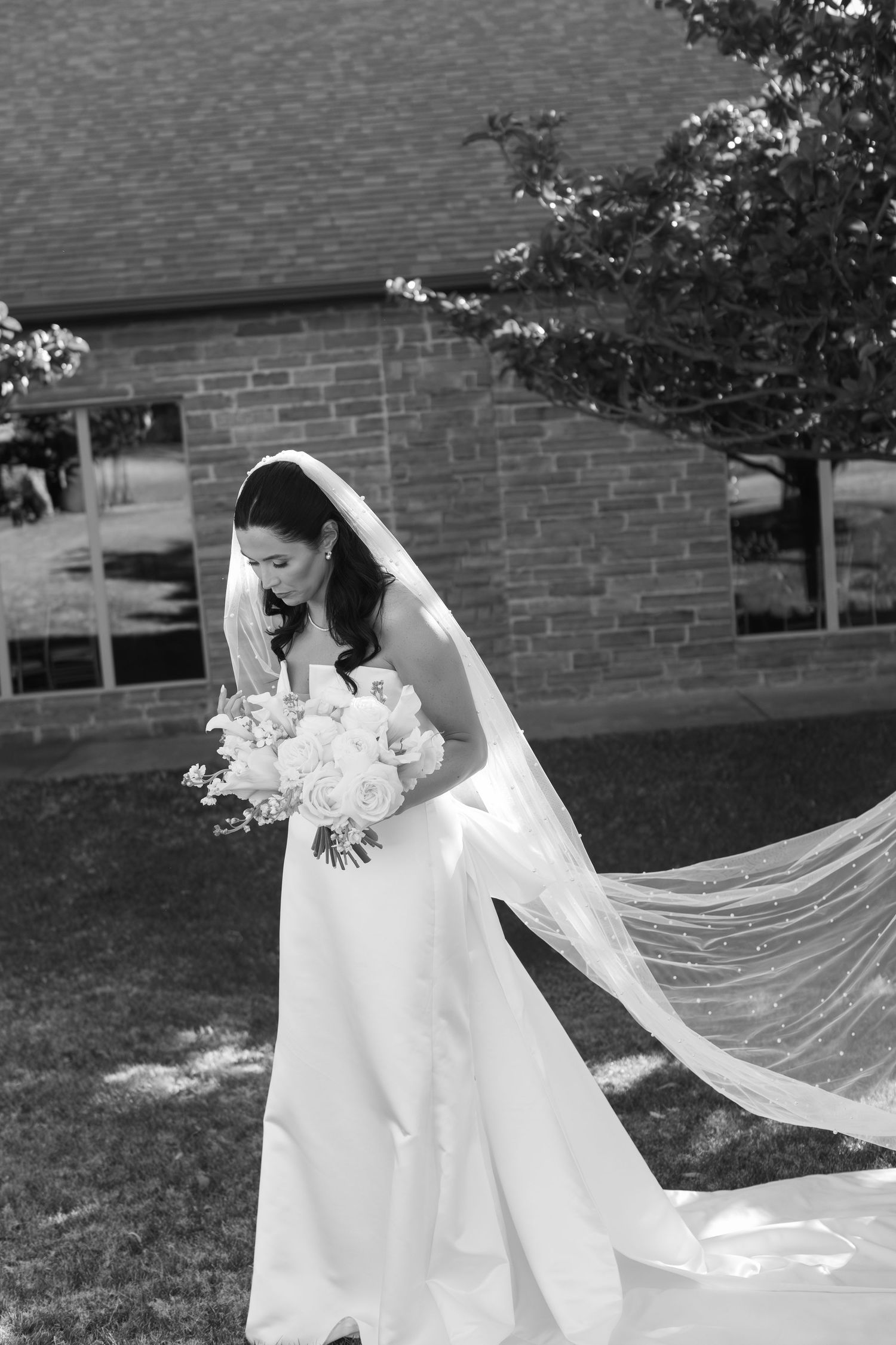 A flowing white wedding veil trails dramatically across a grassy lawn in black and white photography.