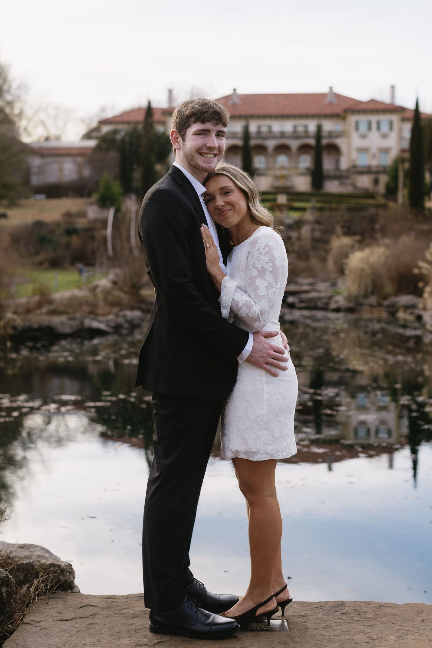 A couple poses for a romantic portrait by a reflective pond with a grand mansion in the background.