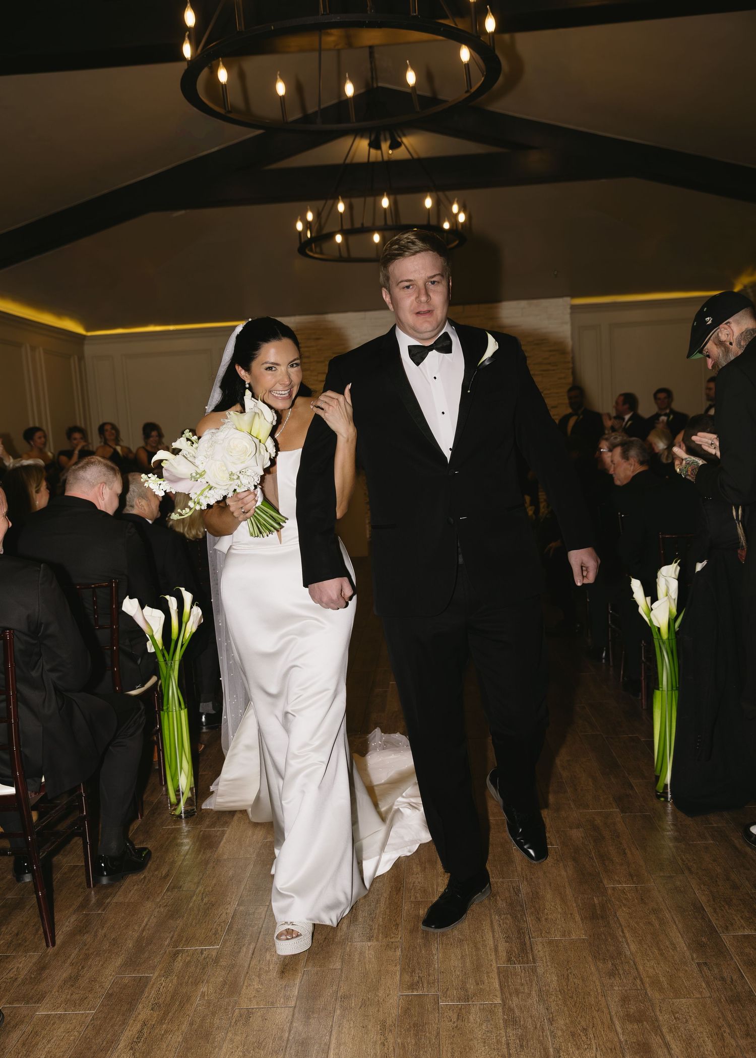 Wedding reception guests dancing on a wooden floor under chandelier lighting.