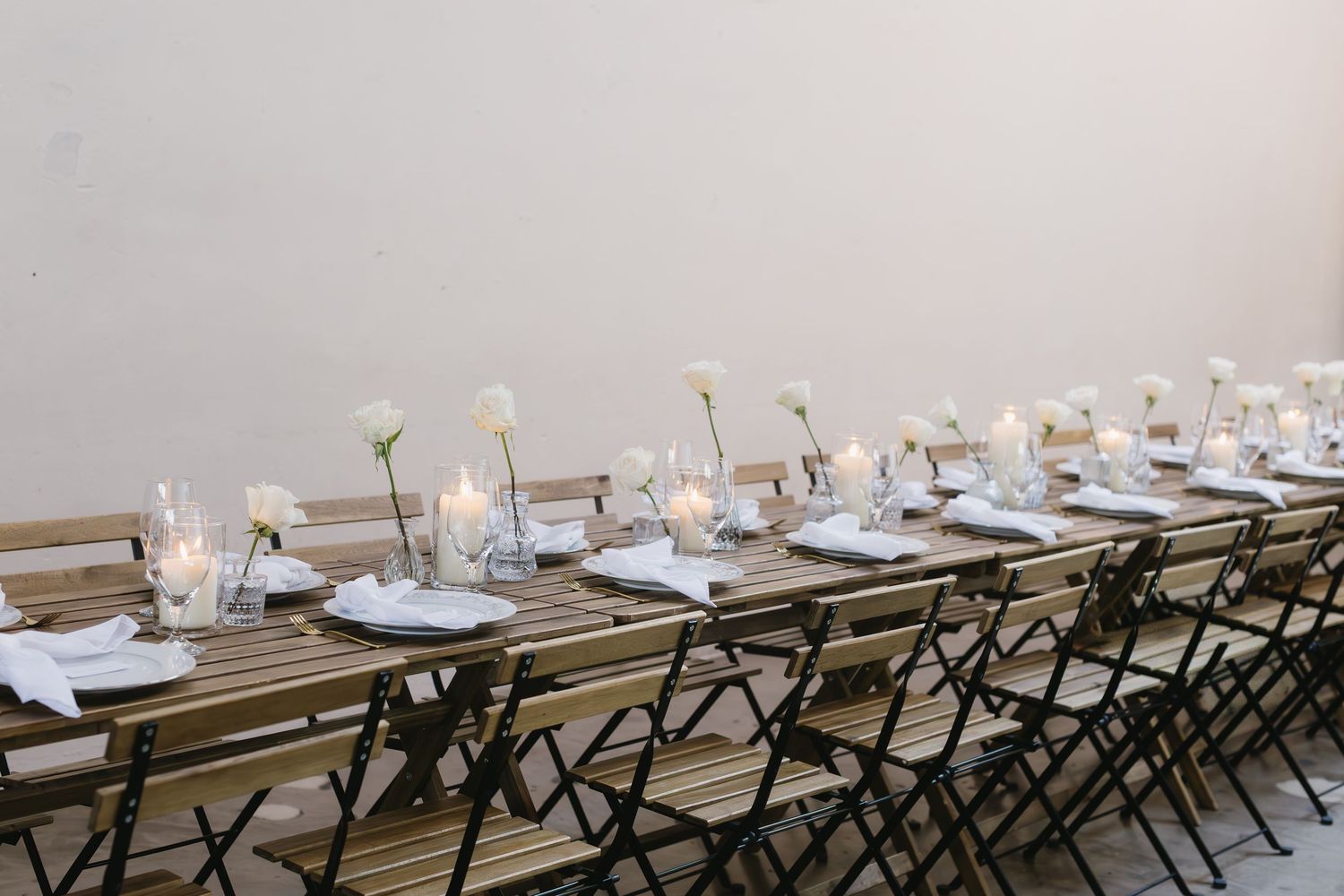 Elegant long wooden dining tables set with white plates and floral centerpieces at an outdoor reception venue.
