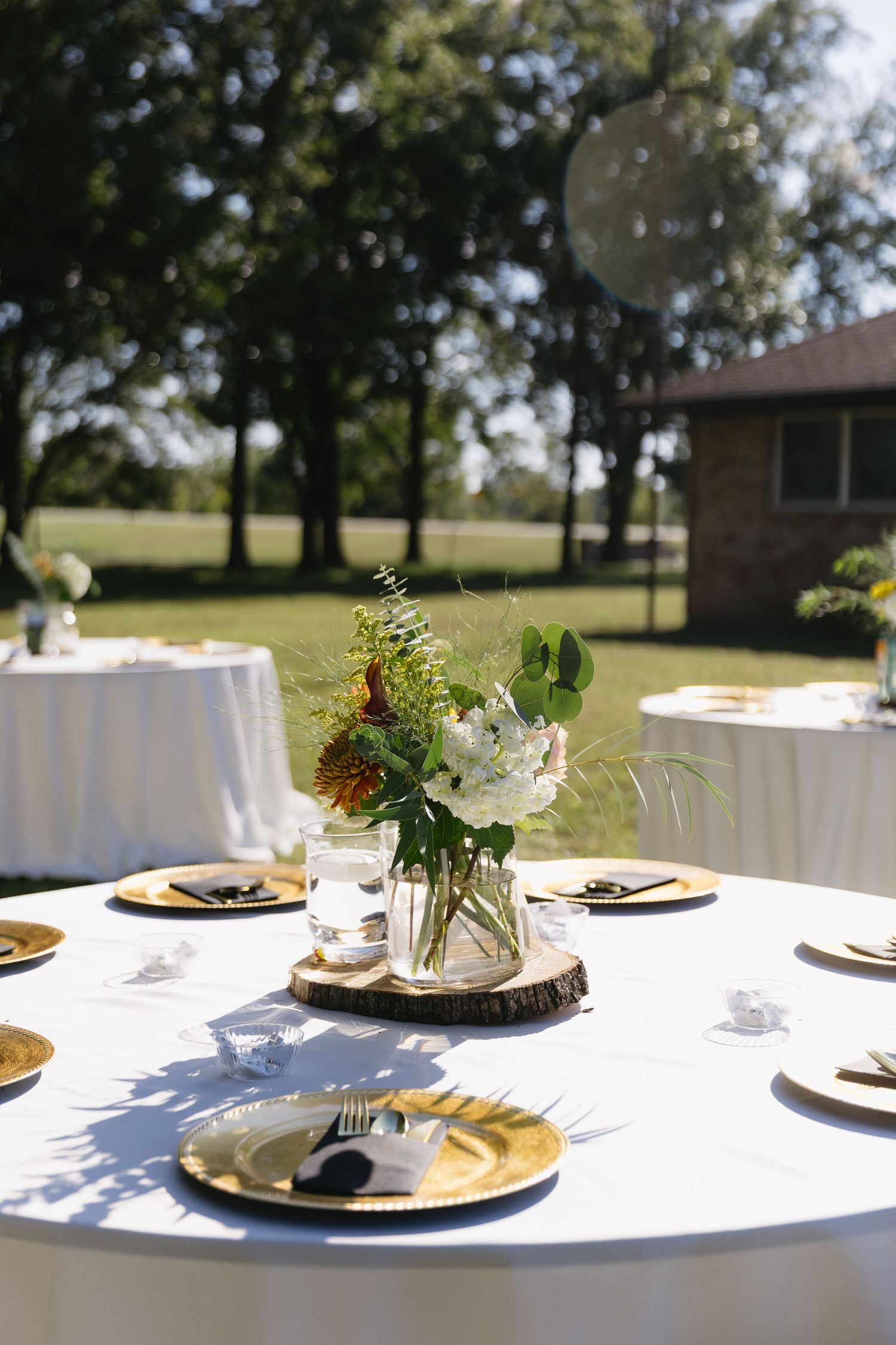 An elegant outdoor wedding reception table set with white linens, gold plates and mason jar centerpieces.