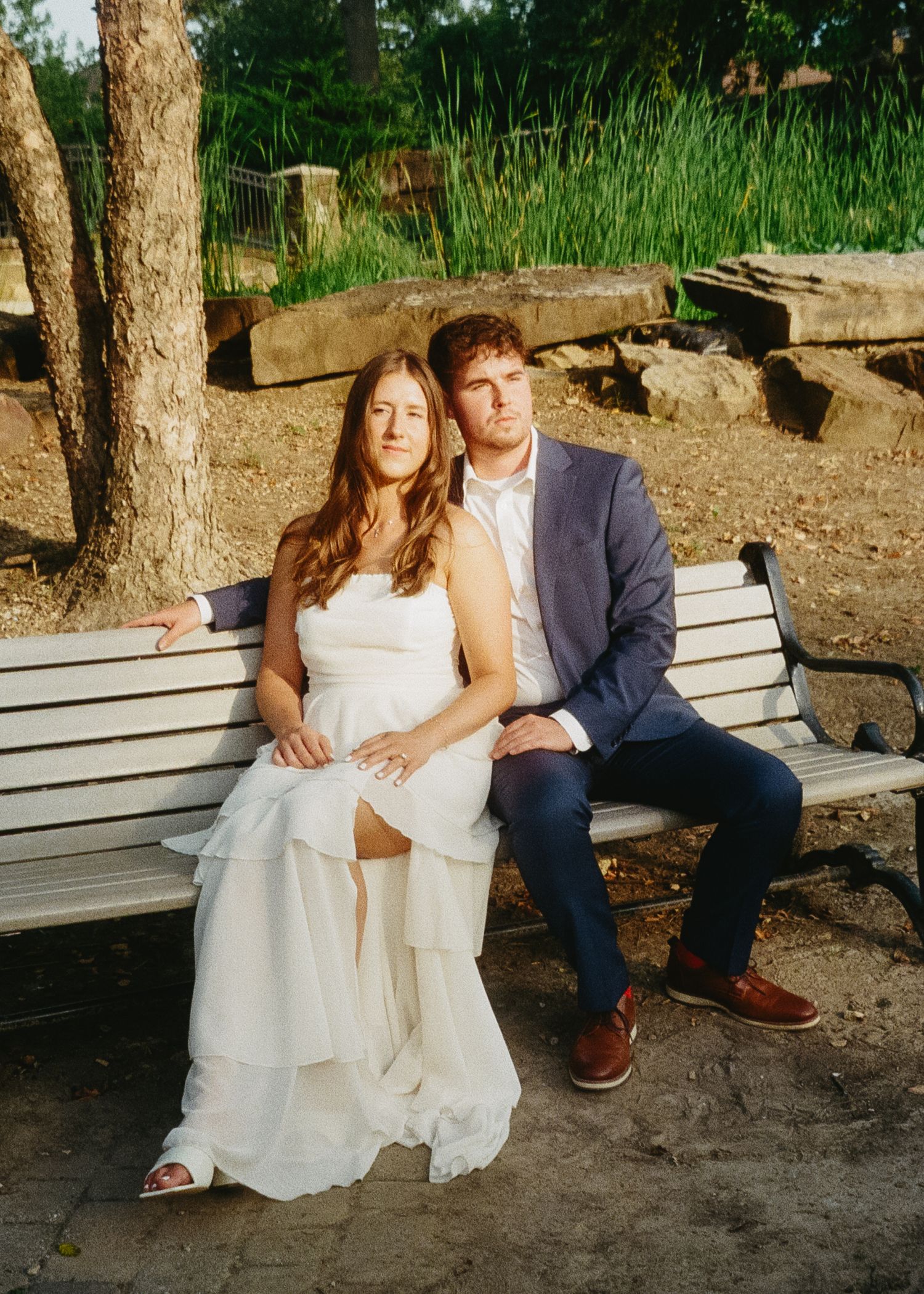 A couple sits together on a wooden park bench surrounded by trees in a romantic outdoor setting.