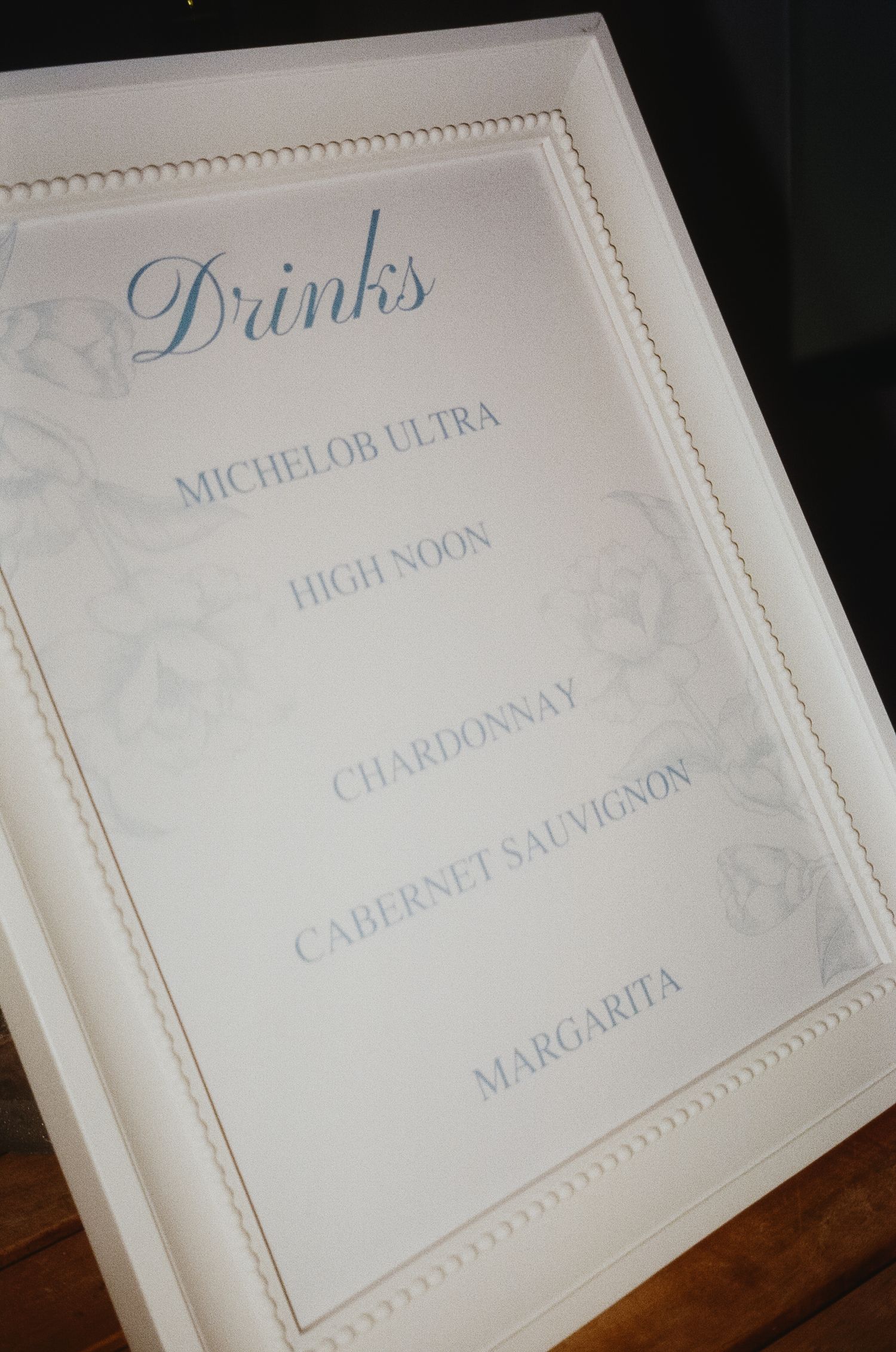 White wedding reception drink menu cards with elegant blue text and decorative borders displayed on wooden surface.