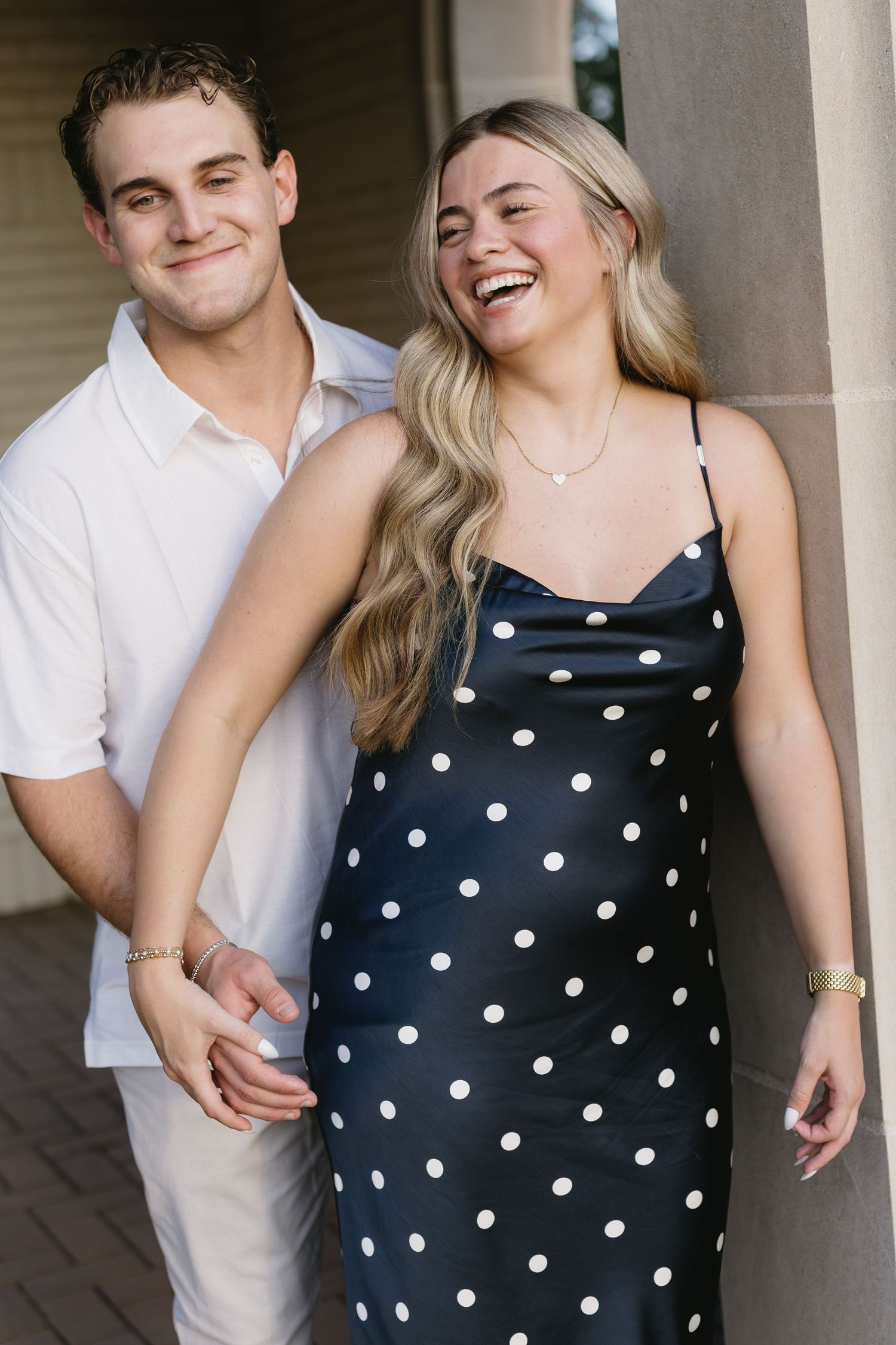 A couple shares laughter while posing in polka dot dress and white shirt under columns.