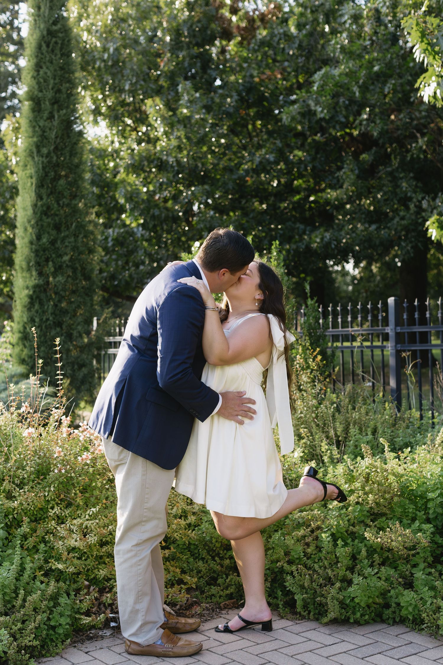 A couple shares intimate moments in a garden setting with lush greenery and iron fencing.