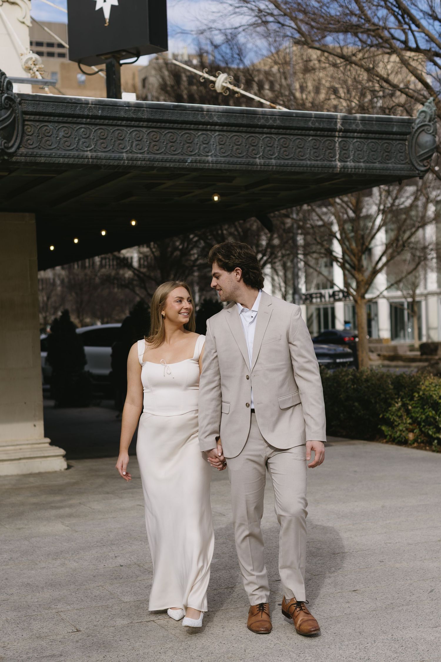 A couple in coordinated formal wear walk hand in hand under an ornate building entrance.