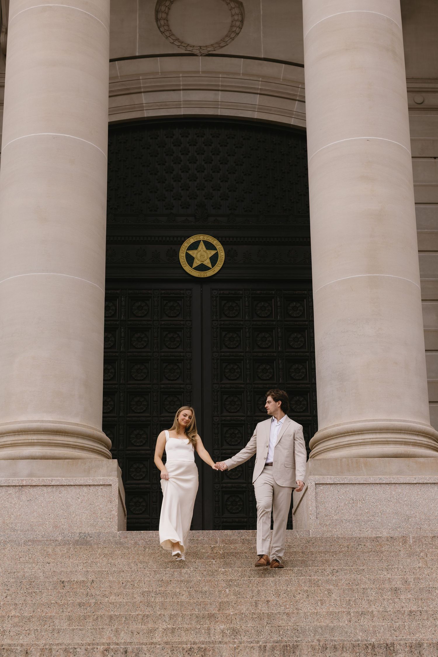 Couple in coordinated white outfits pose playfully on grand stone steps with classical columns.
