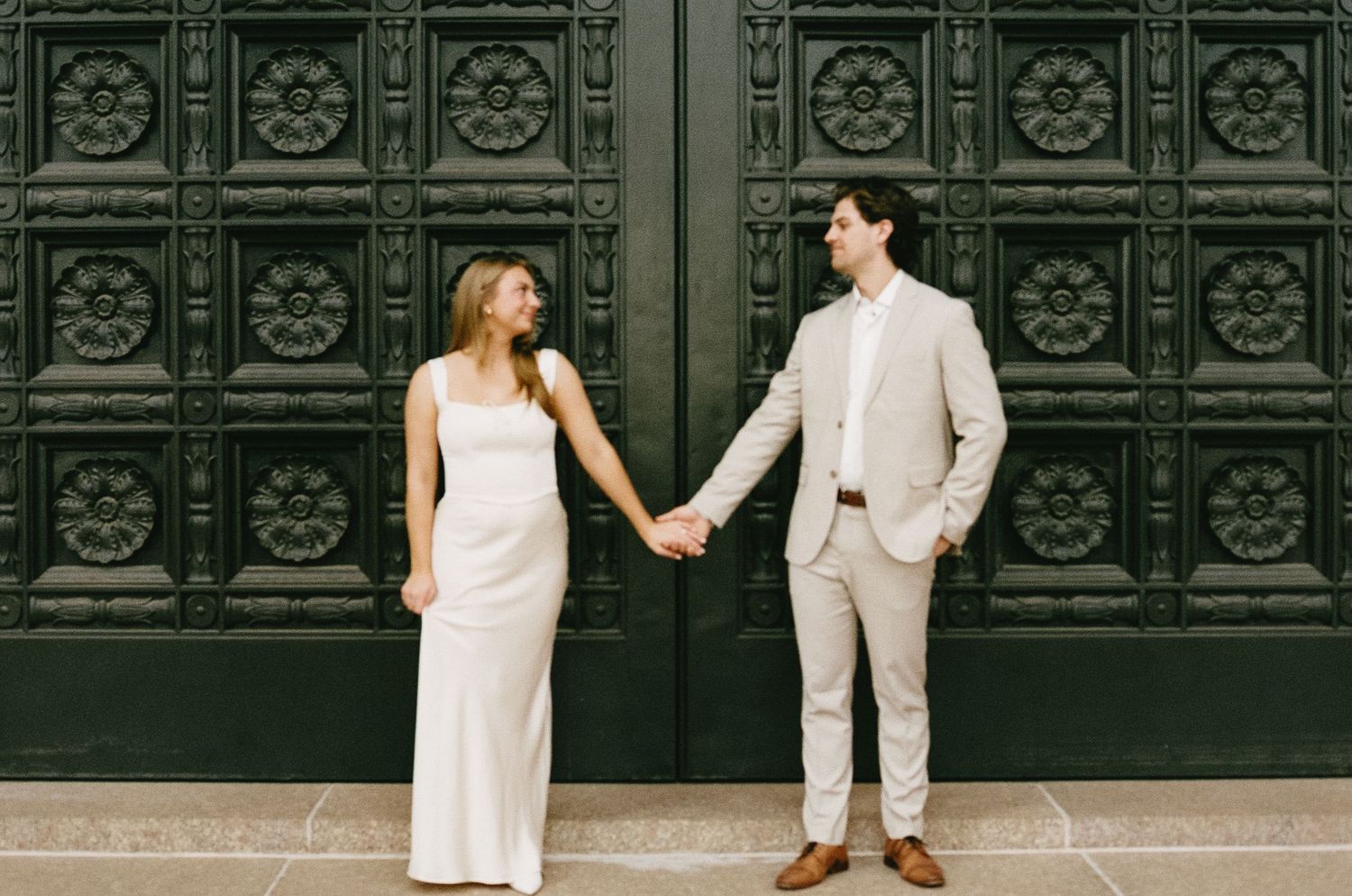 A couple in elegant attire poses playfully against decorative black doors of a historic building.
