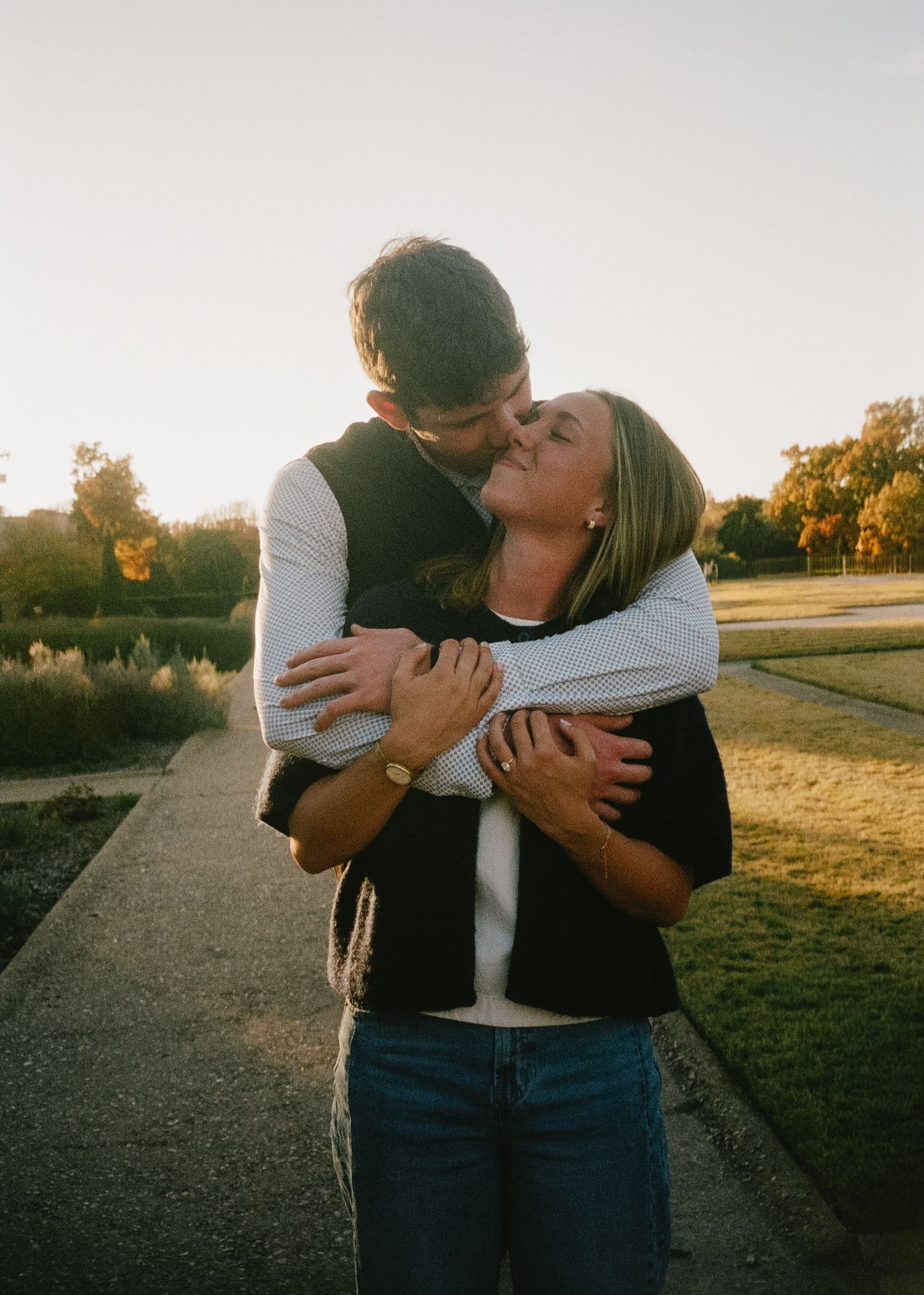 A couple embraces on a pathway at sunset with golden light illuminating the romantic scene in a park setting.