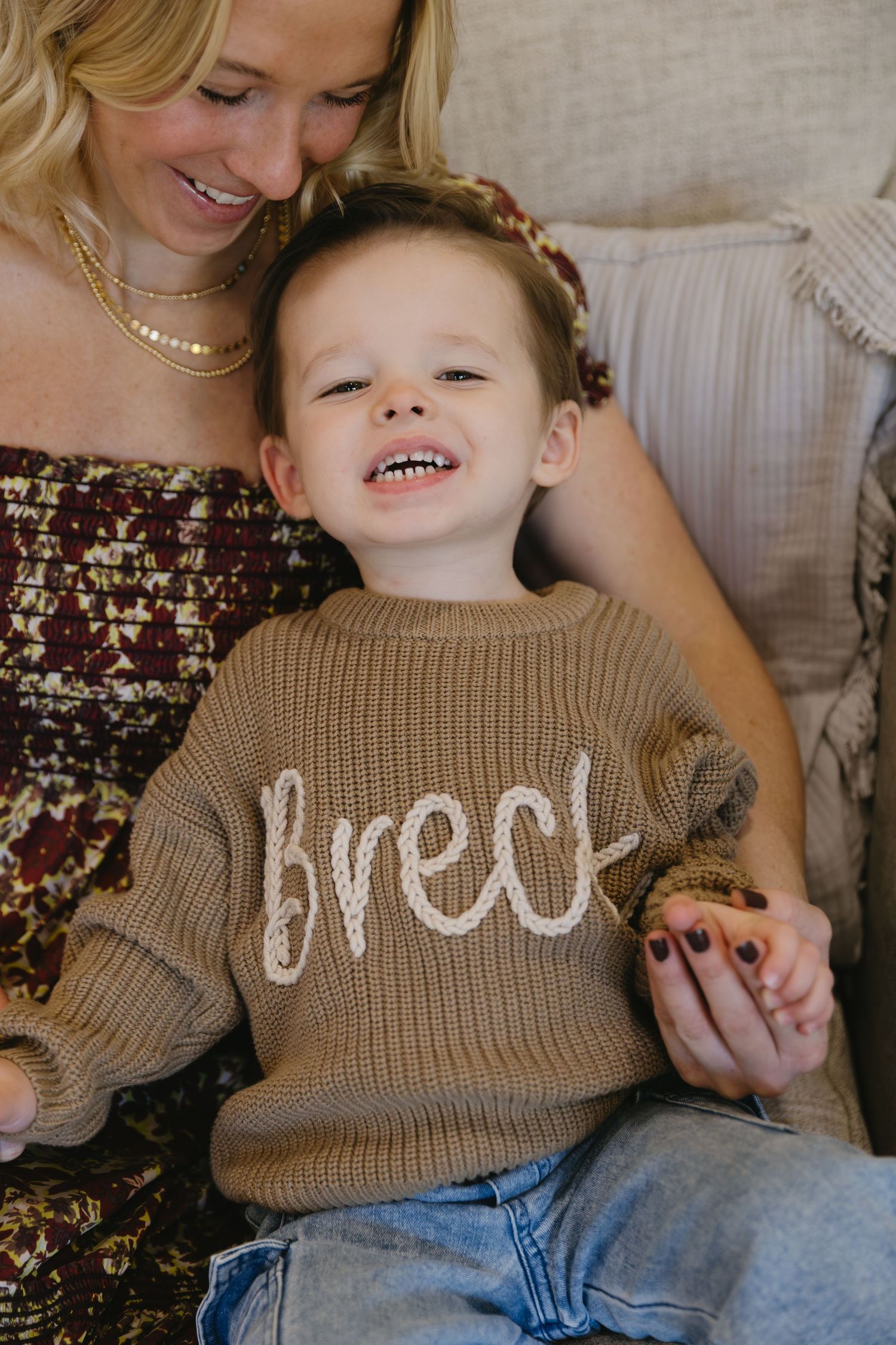 A child wearing a tan knit sweater smiles joyfully while sitting with someone in a floral dress.