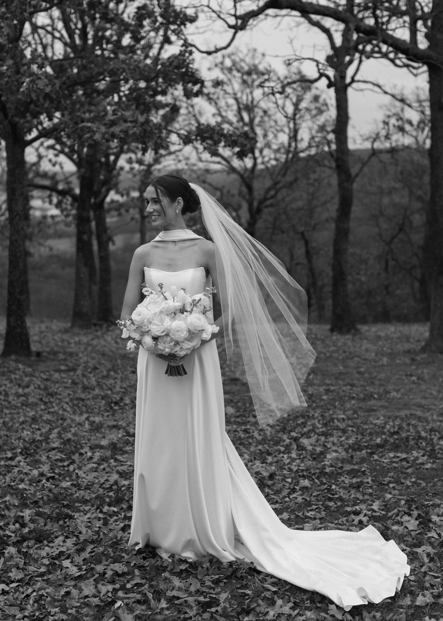 Black and white wedding photograph showing flowing dress and veil against autumn landscape.