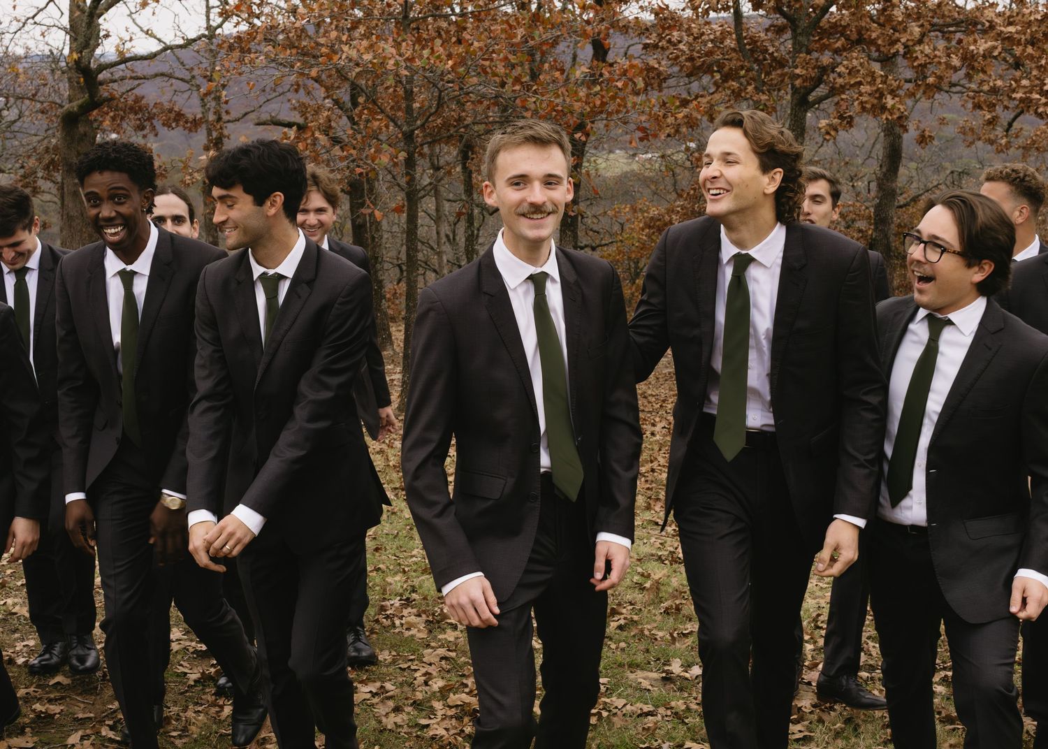 Wedding party in black suits and ties standing outdoors among autumn trees.