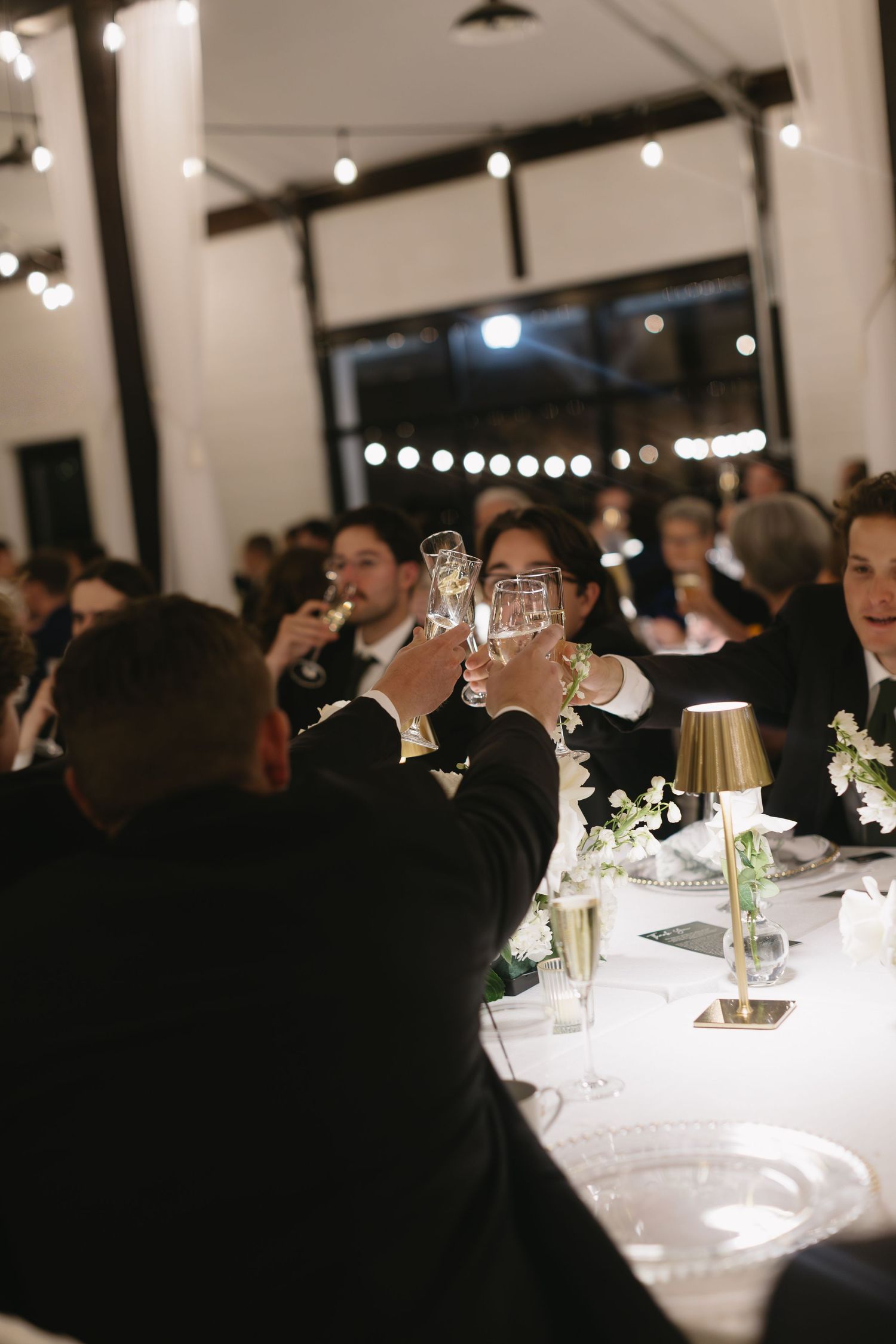 Wedding reception dinner setup with string lights and white tablecloths in an indoor venue.