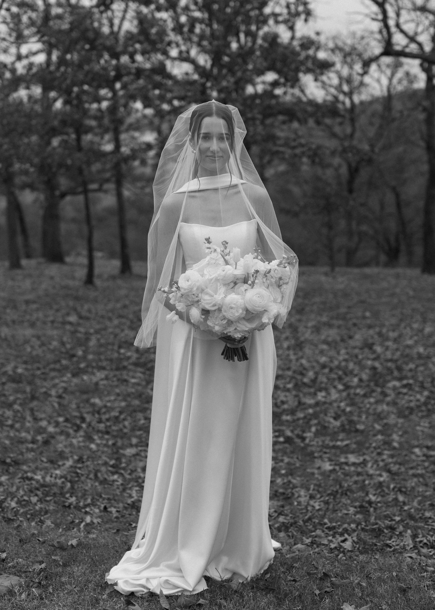 Black and white photo of bride holding white bouquet among fallen leaves.