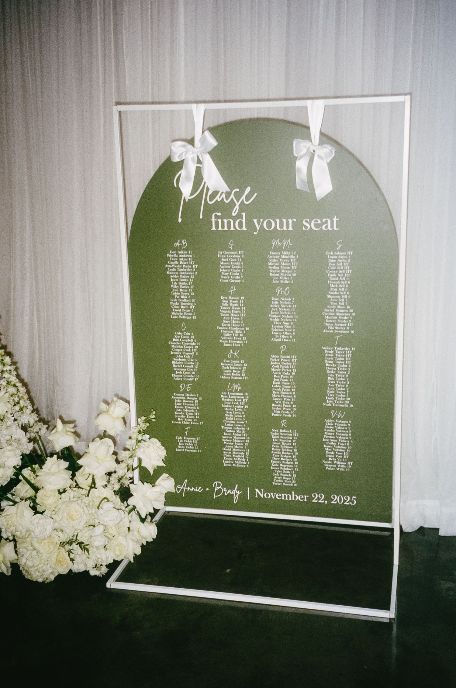 Wedding seating chart display with white text on green panels decorated with white flowers.