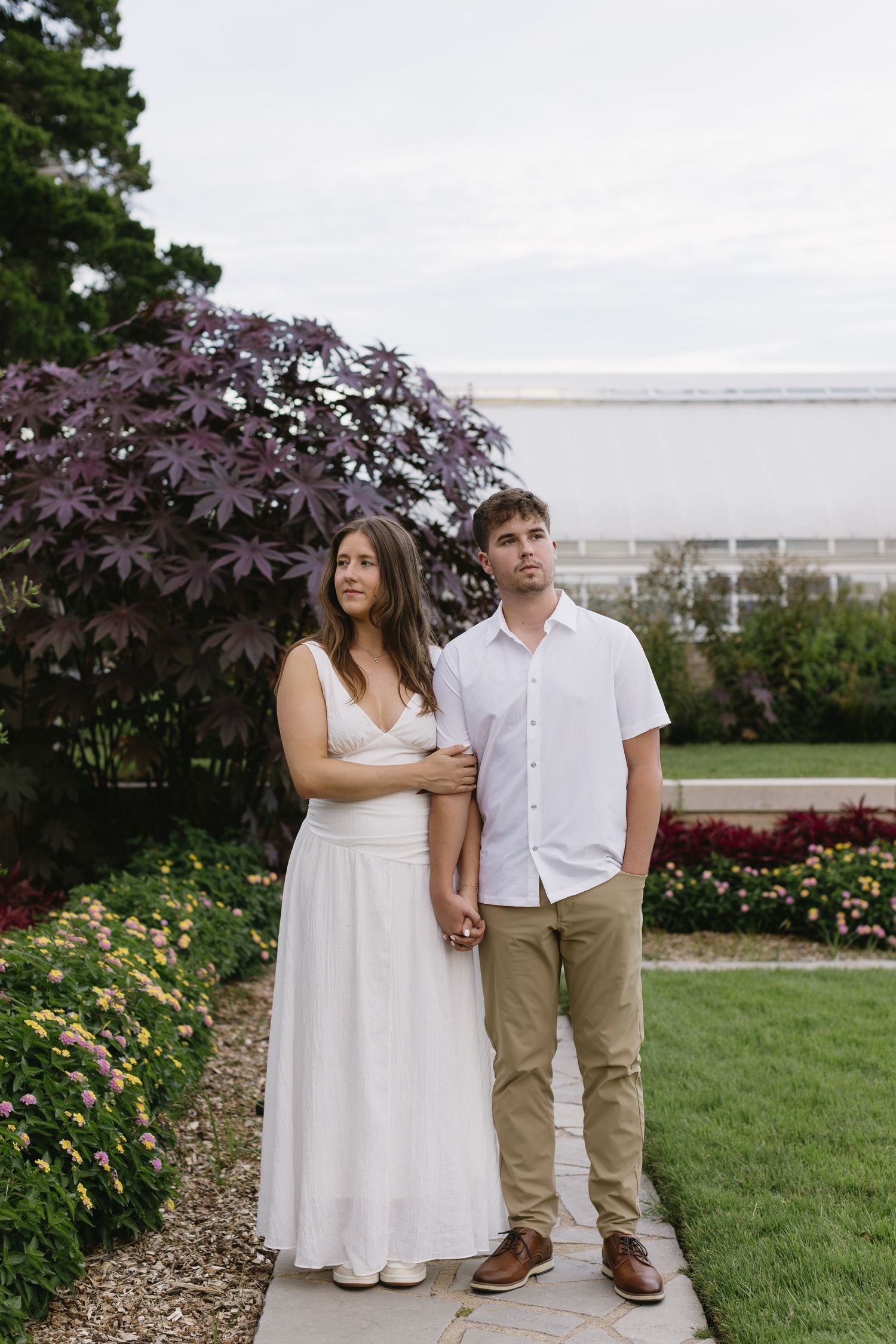 A couple poses for a portrait in a garden with purple Japanese maple and colorful flowers.