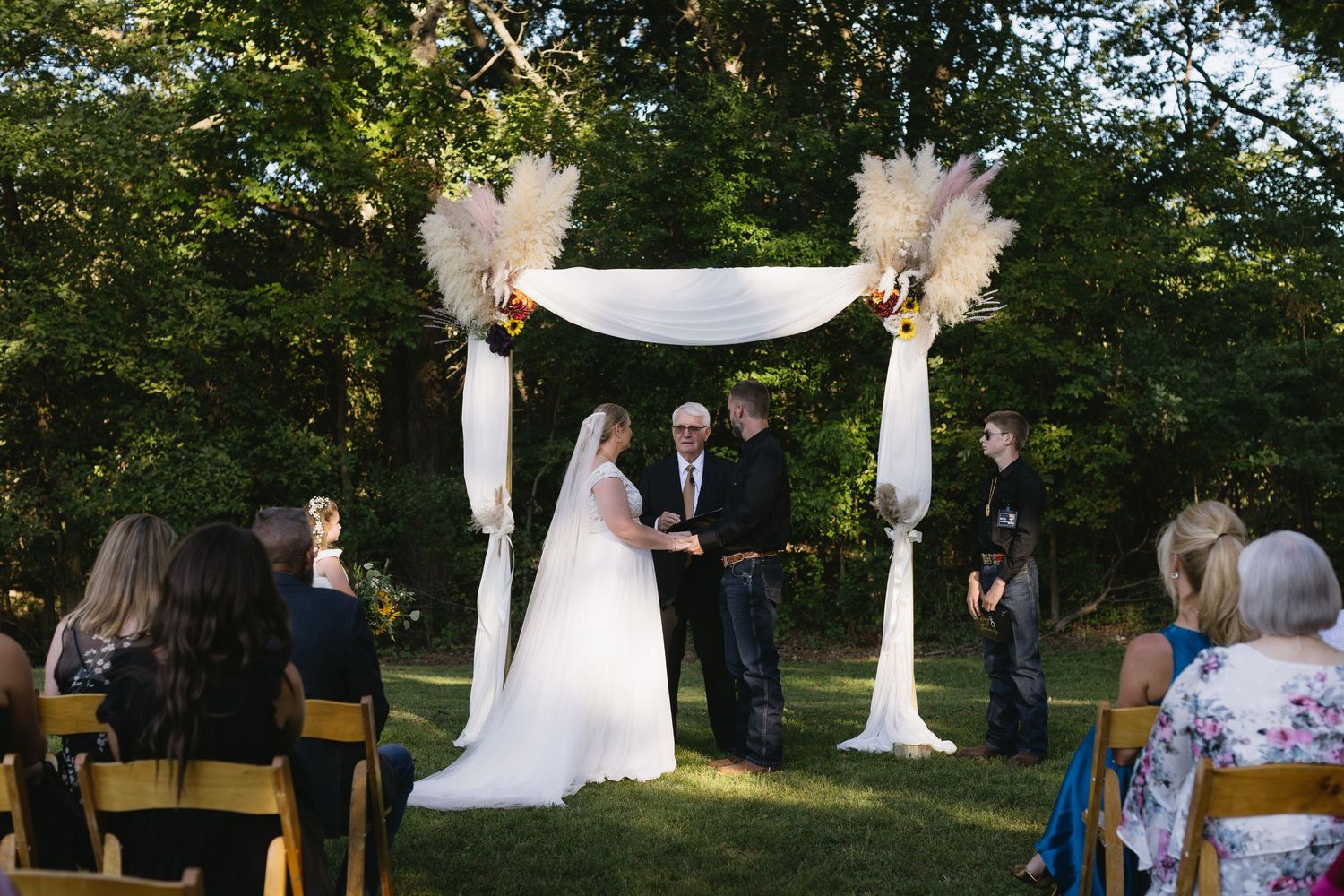 Outdoor wedding ceremony setup with white fabric draped arch and wooden chairs under trees.