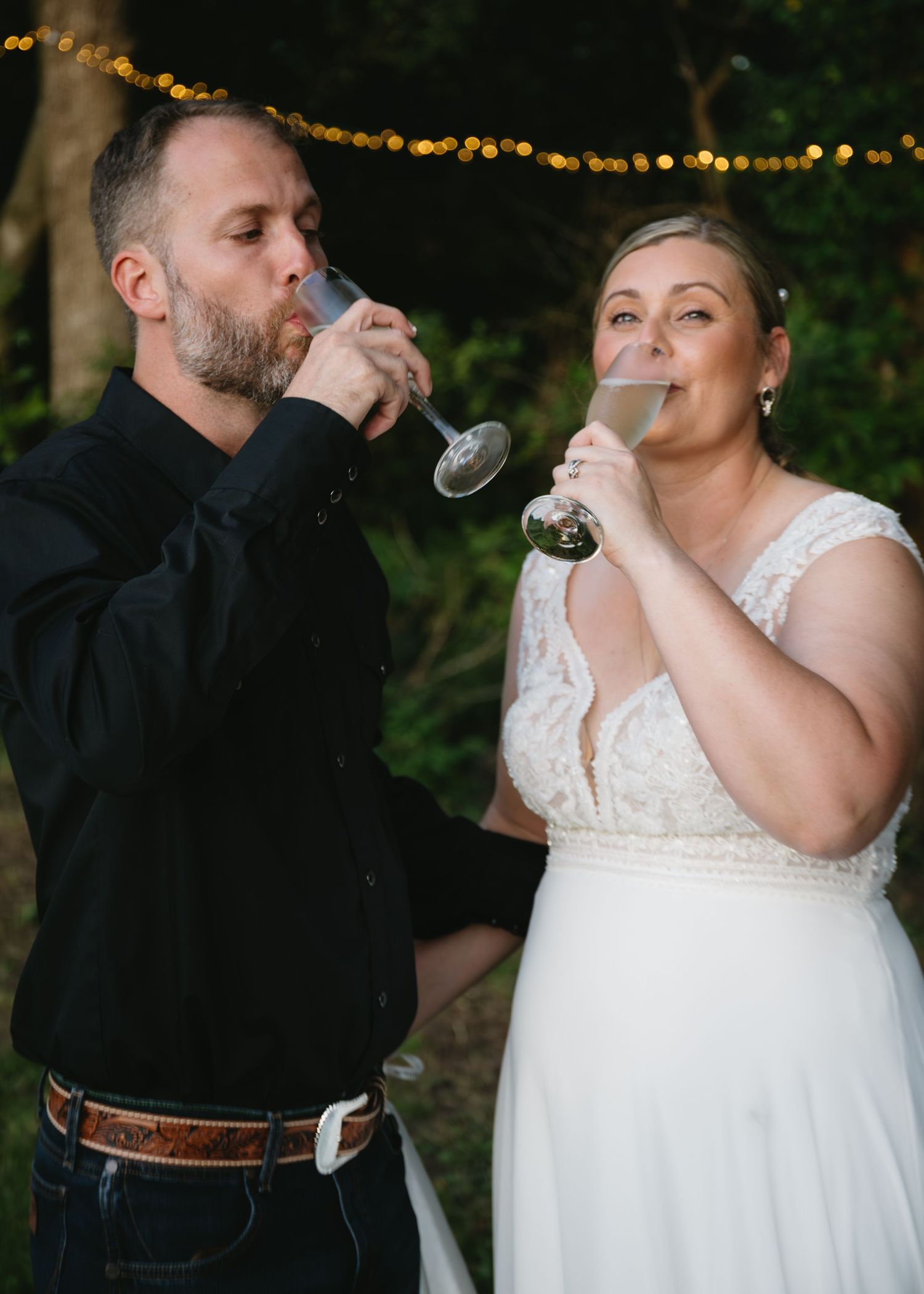 Wedding couple shares a celebratory toast against a backdrop of twinkling lights at dusk.