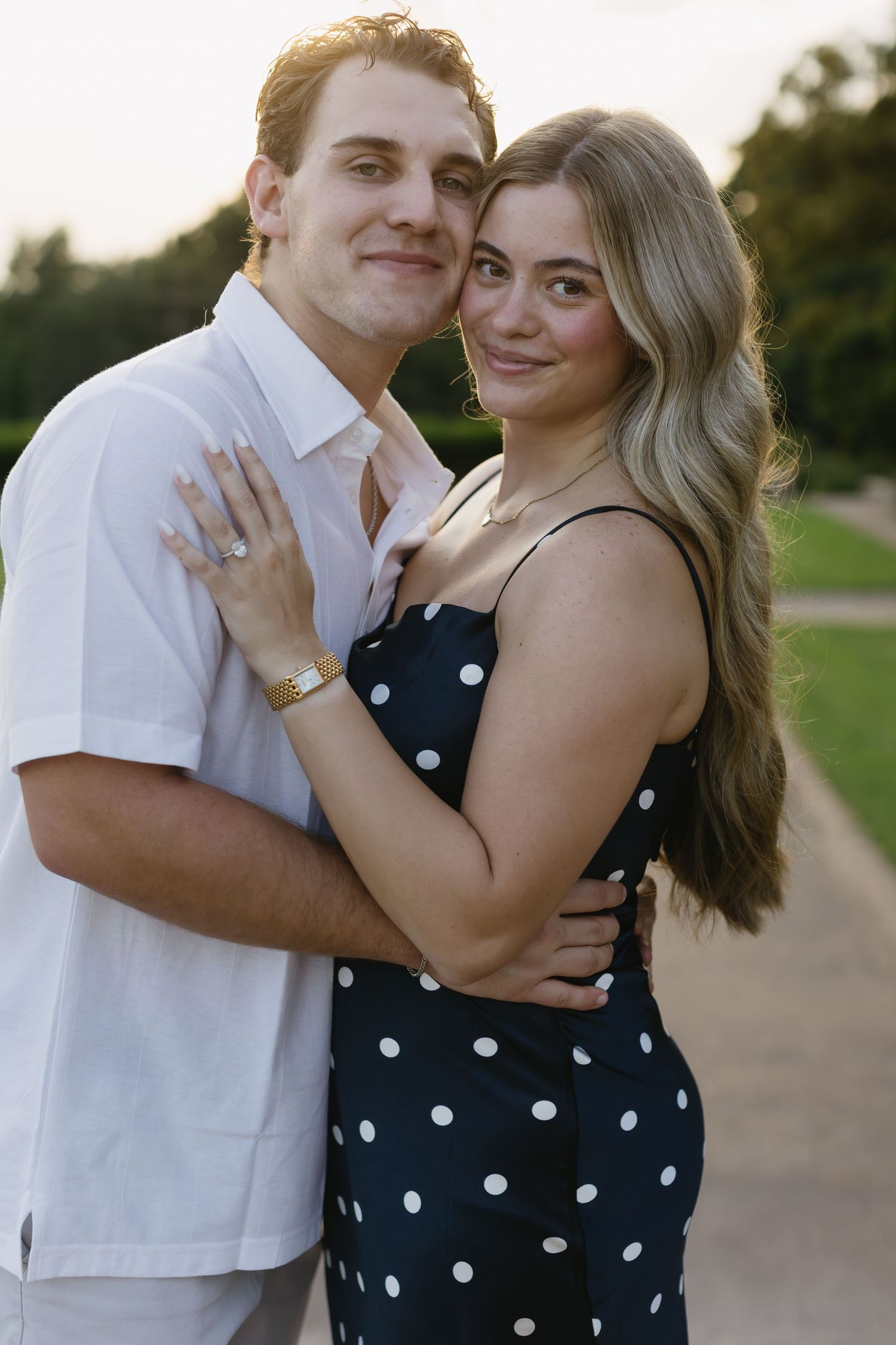 A couple embraces outdoors at sunset, with her wearing a polka dot dress and him in a white shirt.