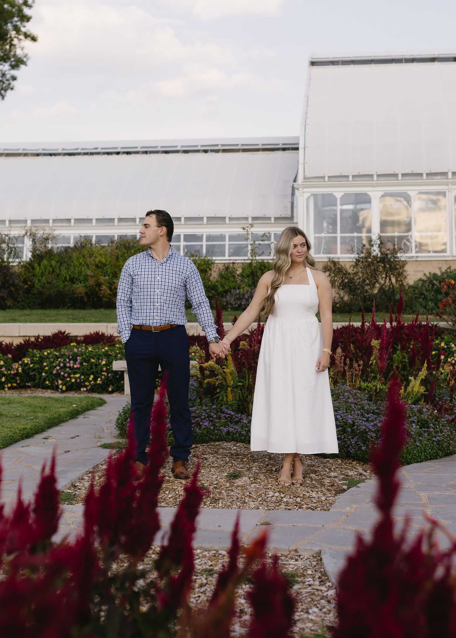 Couple holds hands while walking through a garden with vibrant red flowers and a white building in background.