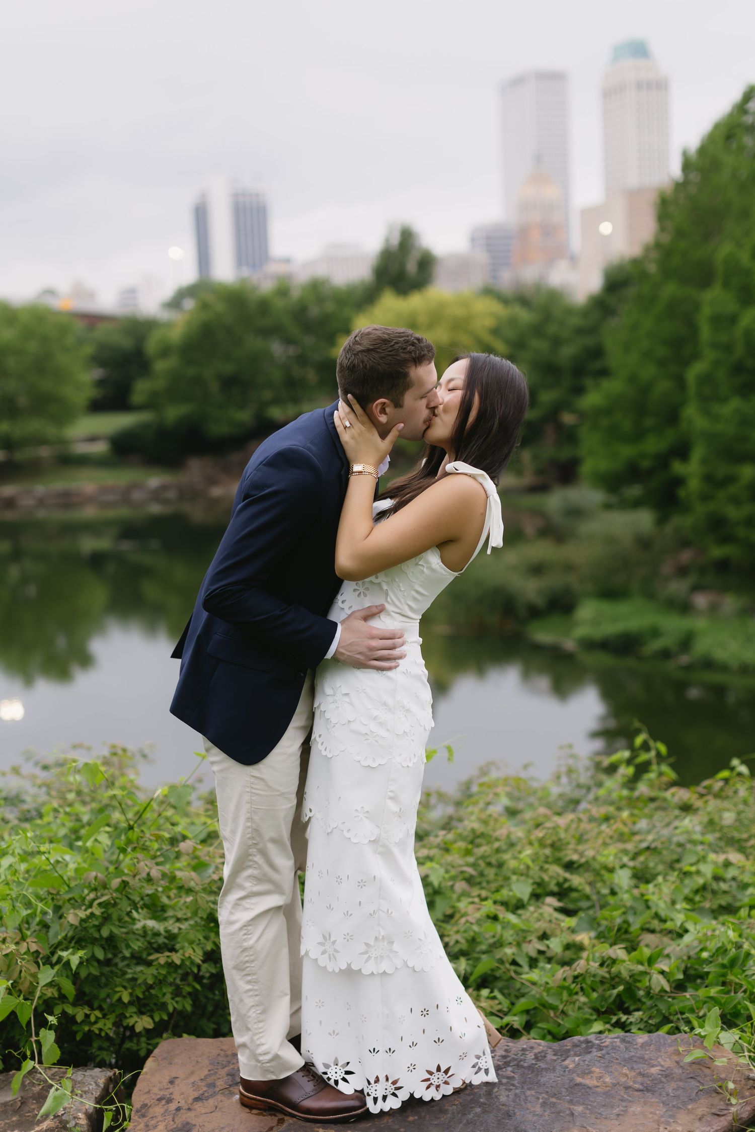 A romantic couple shares a kiss by a scenic pond with city buildings visible in the background.