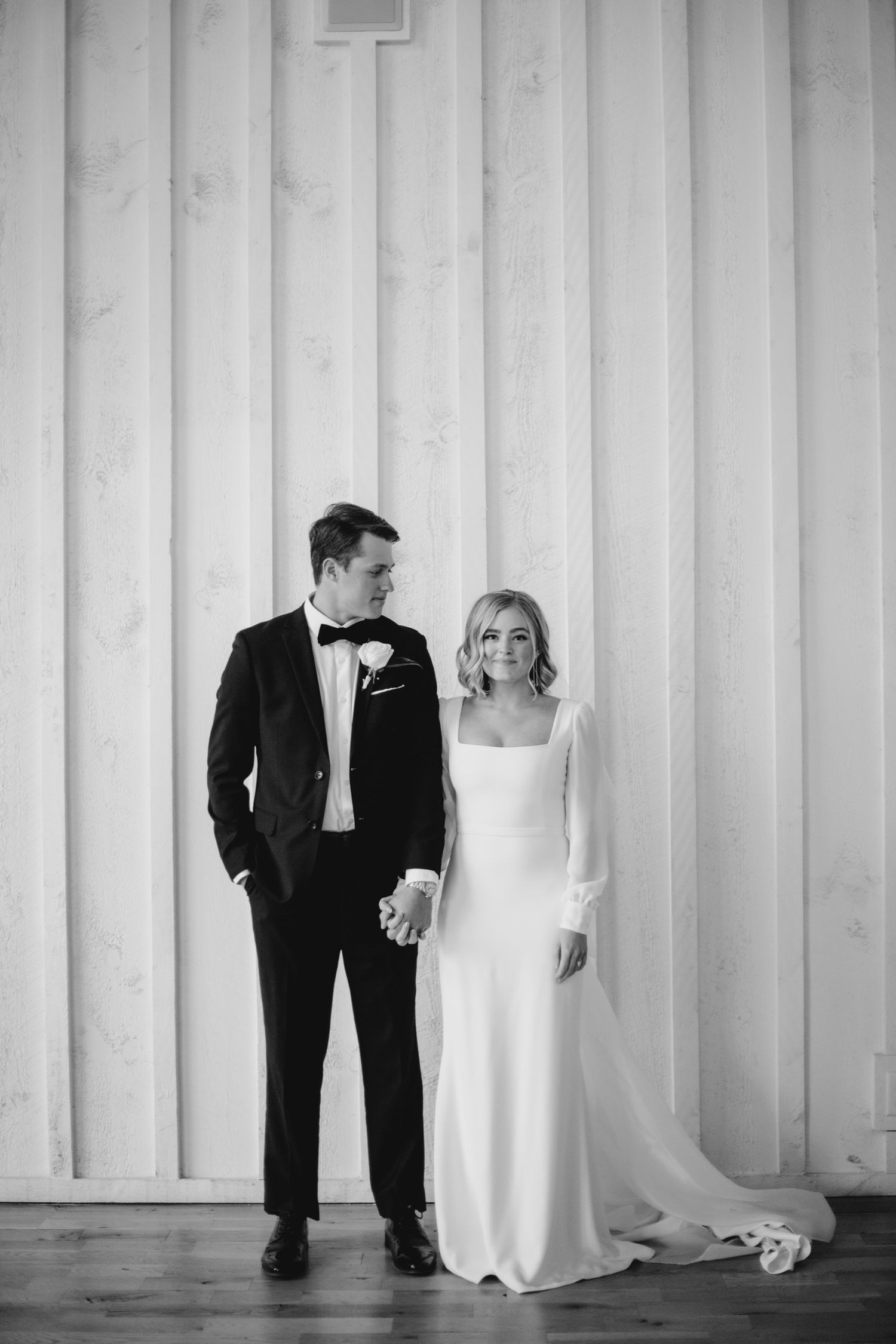 Black and white wedding portrait with bride and groom playfully posing against white striped wall background.